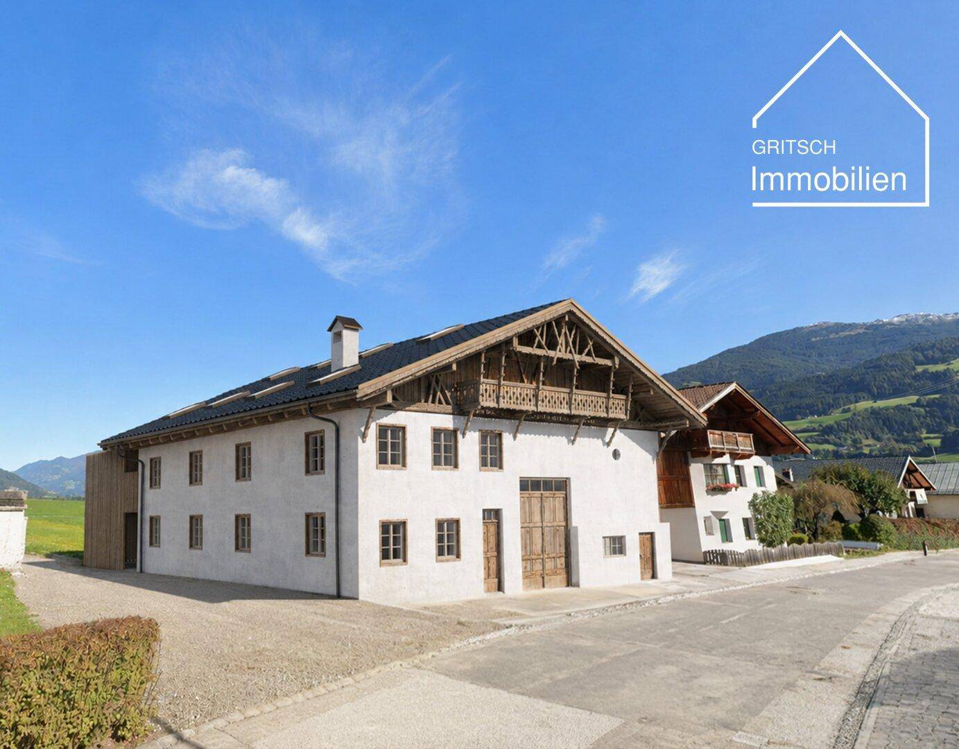 Historisches Gebäude mit weißer Fassade und Holzbalkonen, umgeben von grüner Landschaft und Bergen.