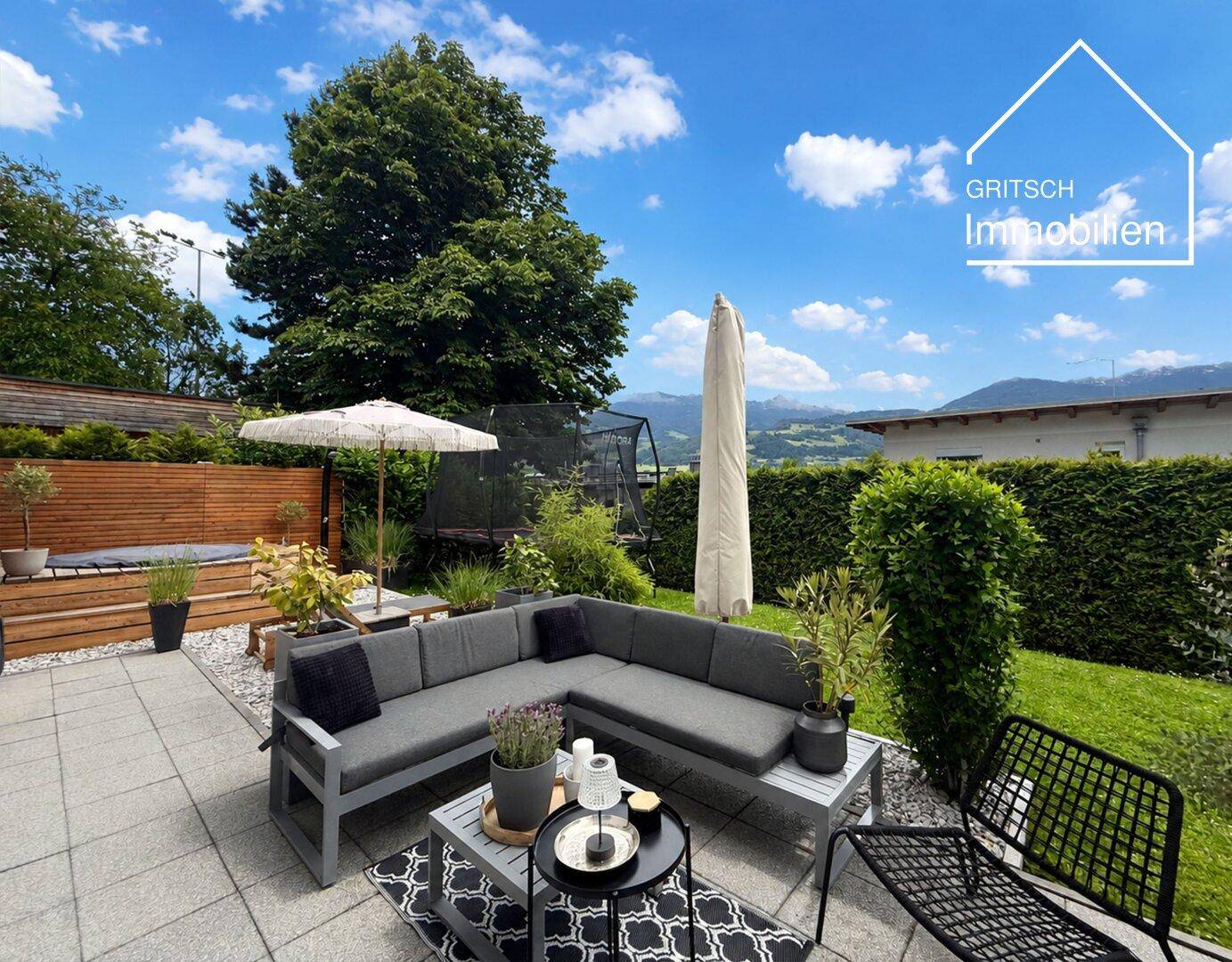 Moderne Terrasse mit Lounge-Möbeln, Sonnenschirm und Garten, umgeben von Hecken und Bäumen.