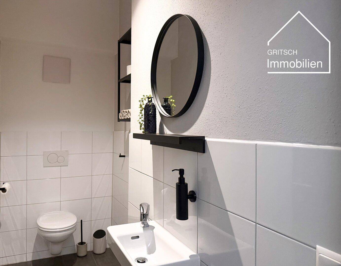 Modernes Badezimmer mit weißen Fliesen, wandhängendem WC und rundem Spiegel.