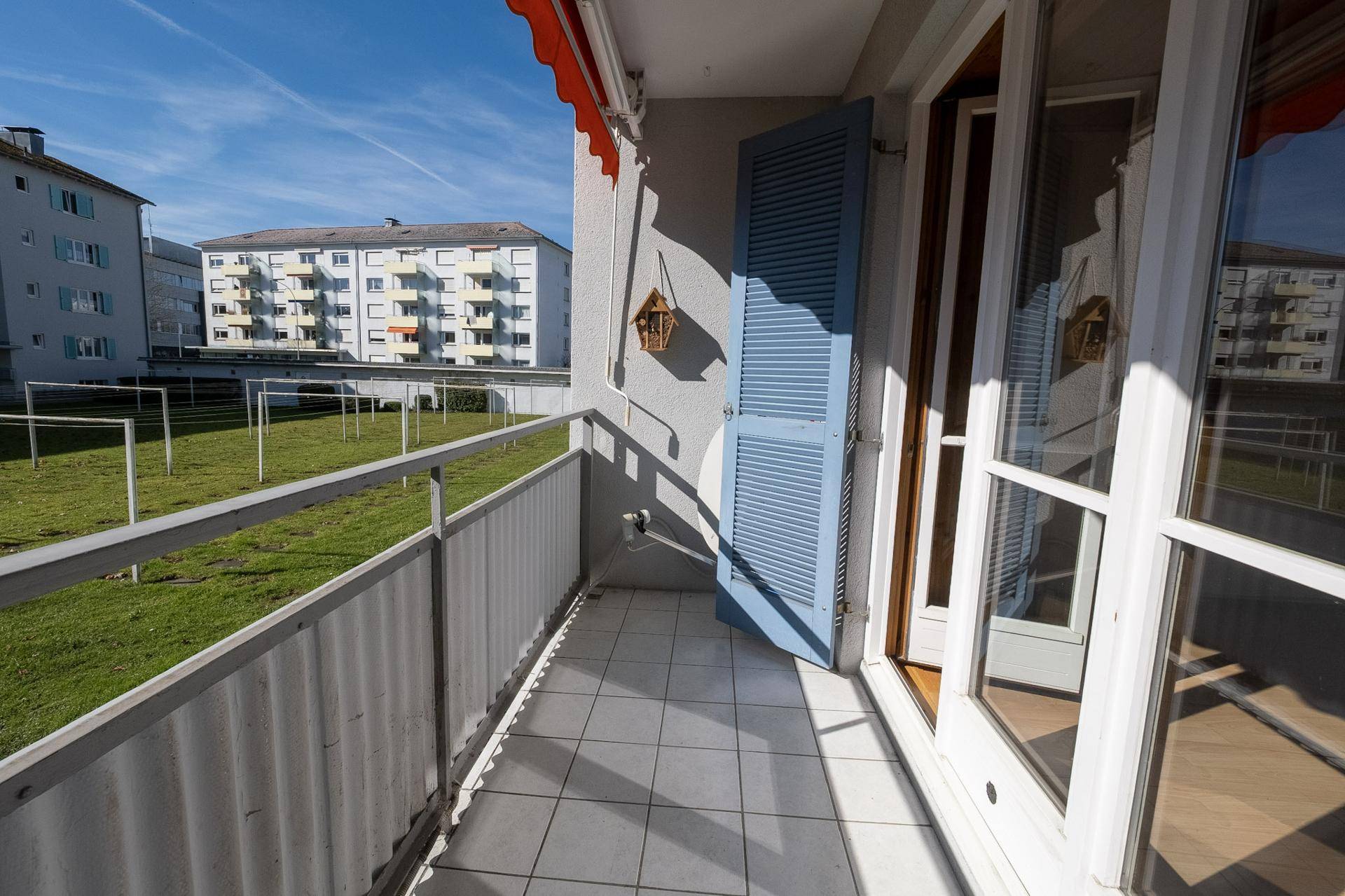 Heller Balkon mit Fliesenboden, Metallgeländer und Blick auf die gemeinschaftliche Grünfläche.