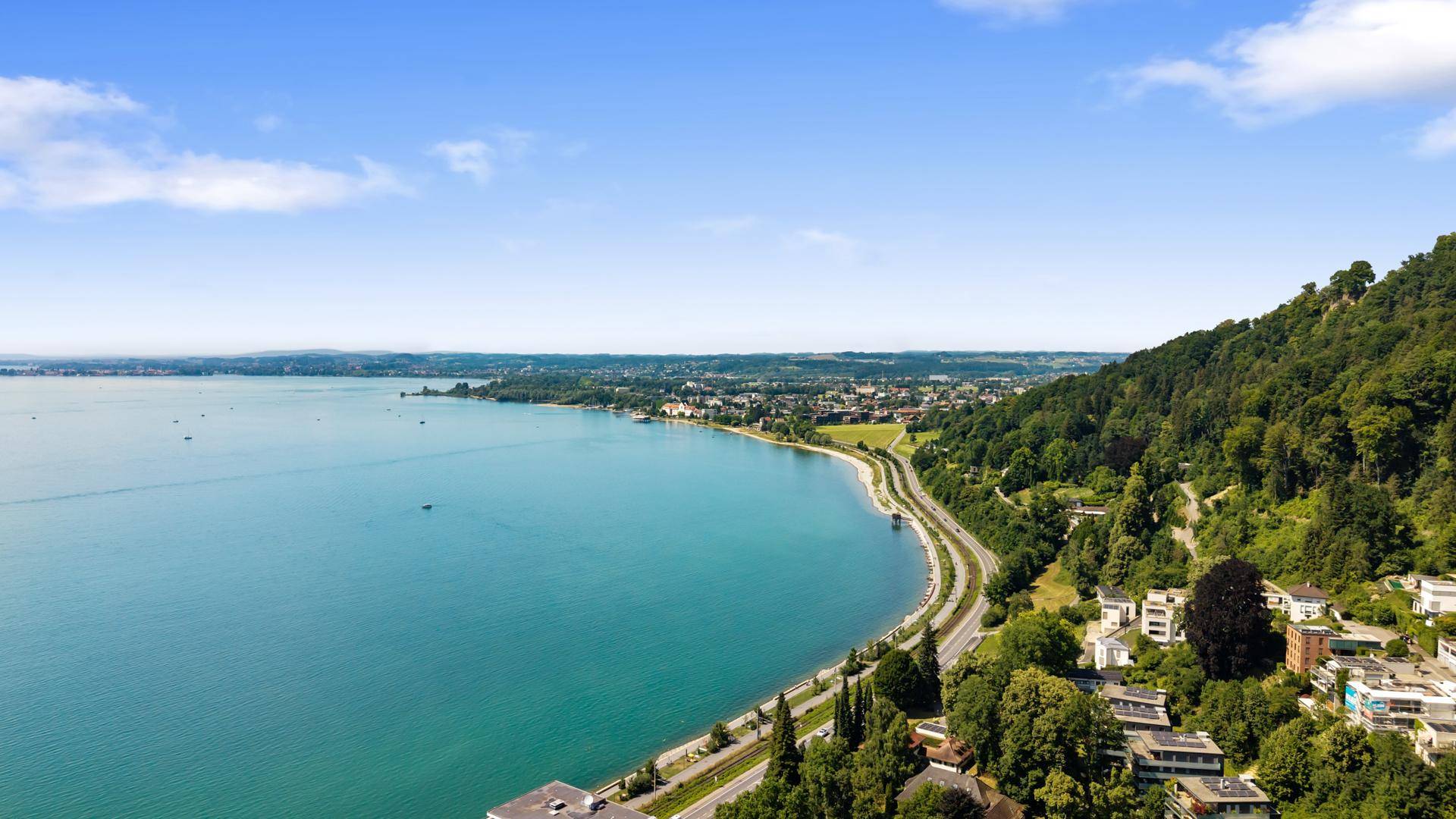 Weitläufiger Blick auf den See mit bewaldeten Hügeln und einer Küstenstraße.