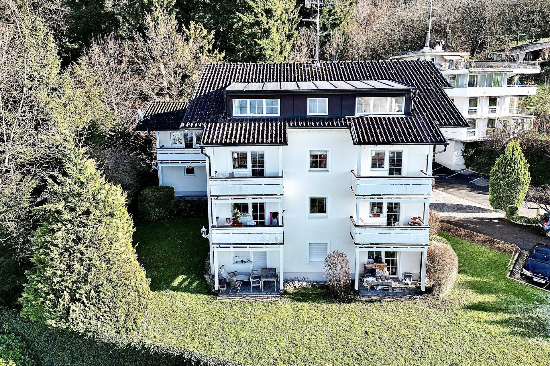 Mehrfamilienhaus mit mehreren Balkonen und Solaranlage auf dem Dach, umgeben von Bäumen.