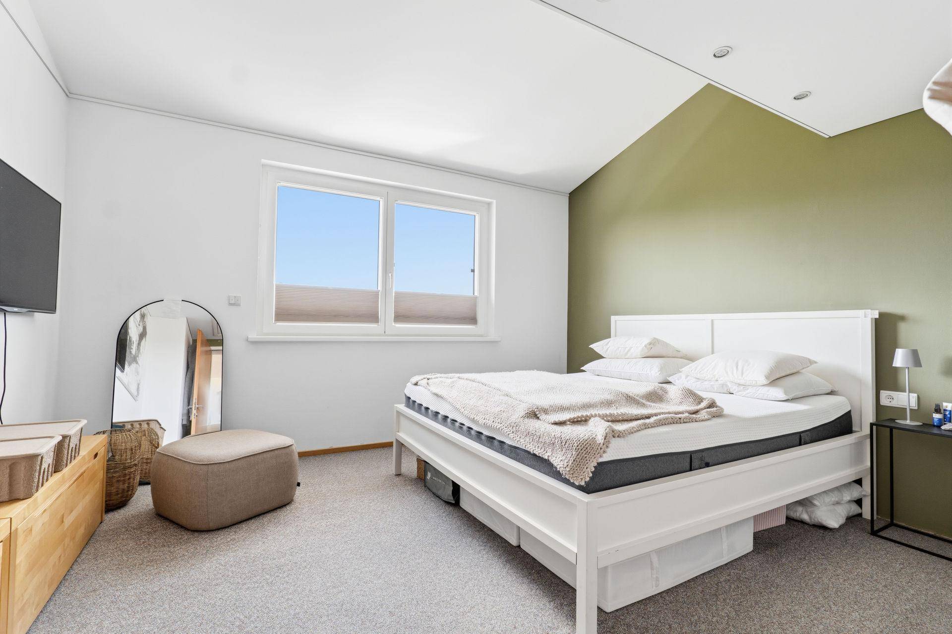 Gemütliches Schlafzimmer mit weißem Bett, grüner Akzentwand und Teppichboden.
