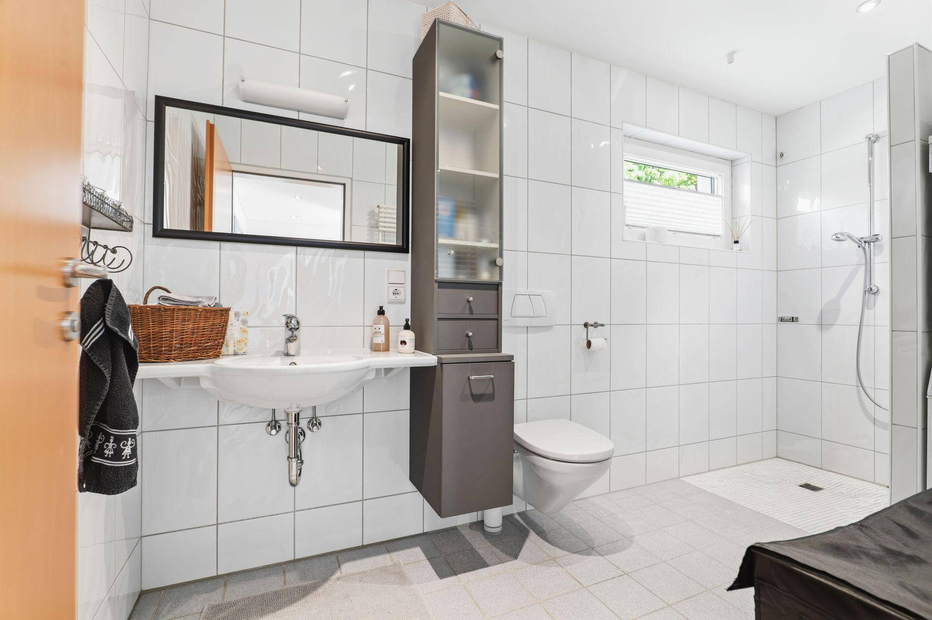 Helles Badezimmer mit weißen Fliesen, Waschbecken, Toilette und Duschbereich, mit Fenster.