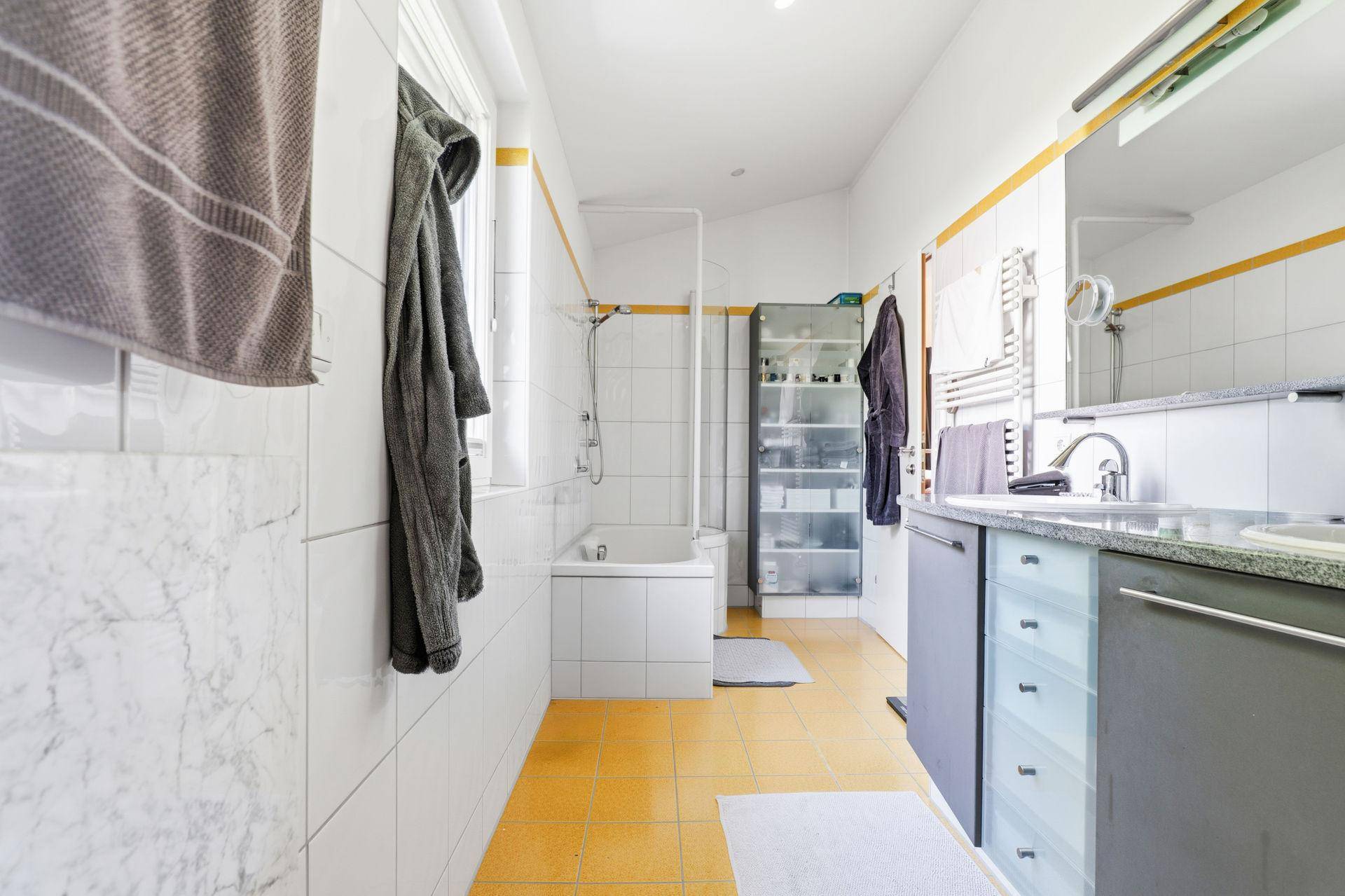 Badezimmer mit gelben Bodenfliesen, Badewanne mit Duschfunktion und Waschbecken.