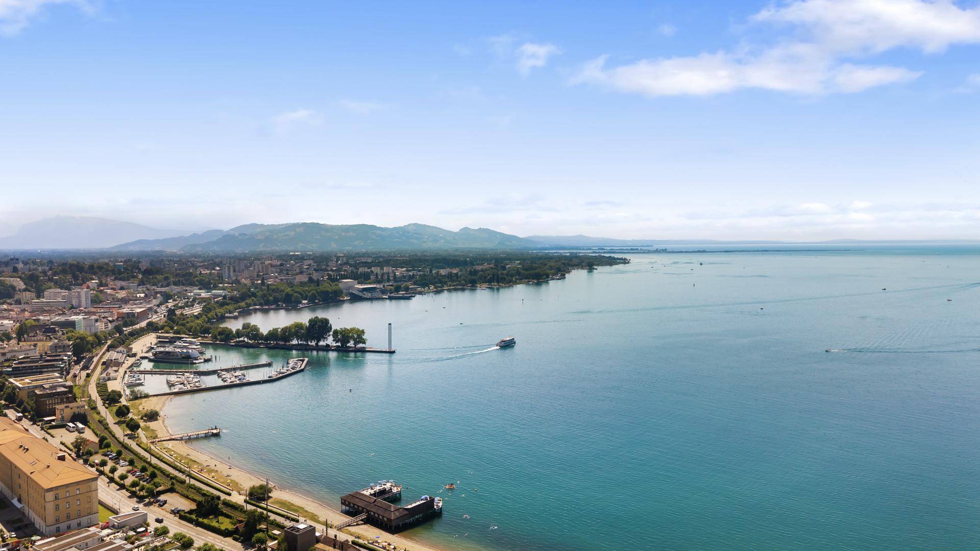 Luftaufnahme des Sees mit Hafen, Stadtgebiet und Bergen im Hintergrund unter blauem Himmel.