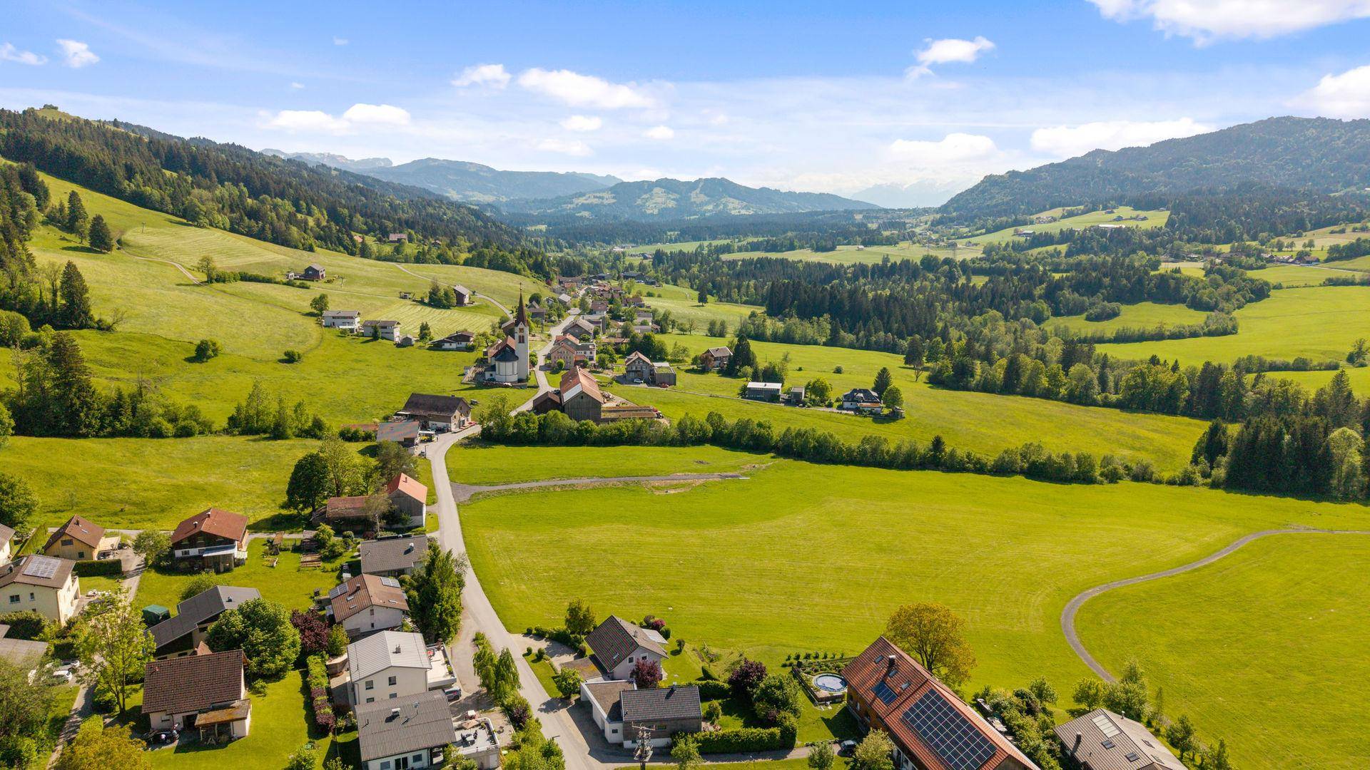 Weitläufige Luftaufnahme eines Tals mit einem Dorf, grünen Feldern und Bergen im Hintergrund.