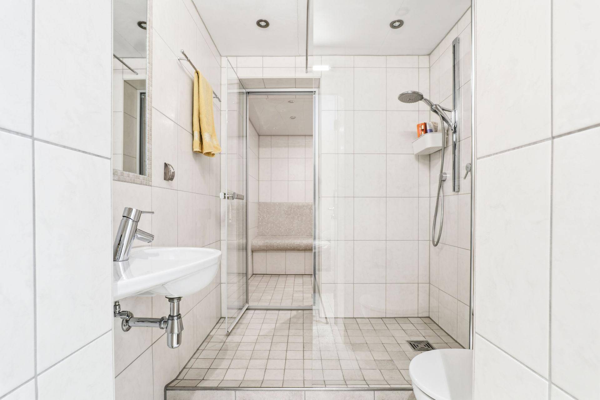 Modernes Badezimmer mit weißen Fliesen, ebenerdiger Dusche und kleinem Waschbecken.