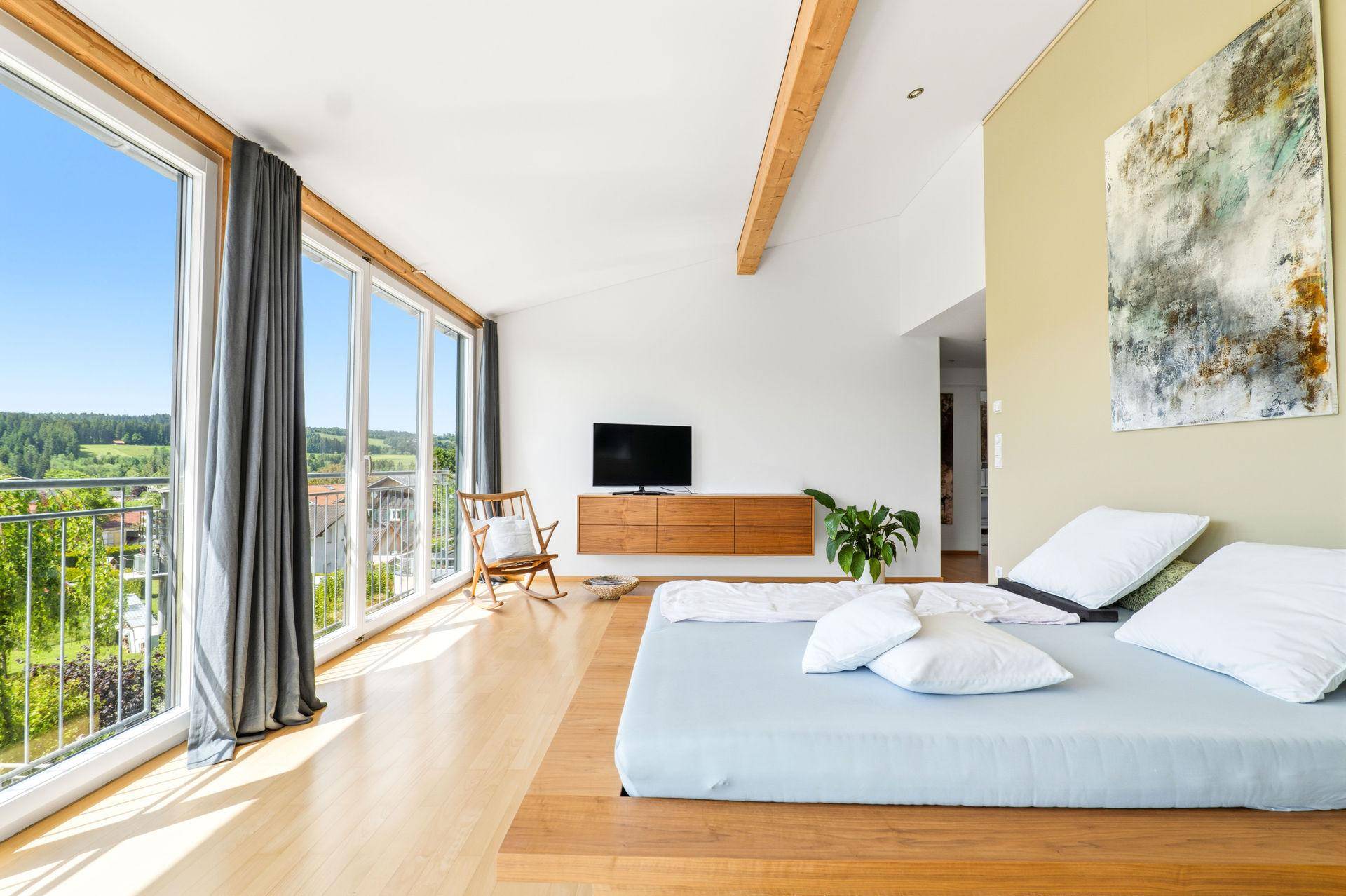 Geräumiges Schlafzimmer mit Holzbett, TV-Möbel und großen Fenstern mit Blick ins Grüne.