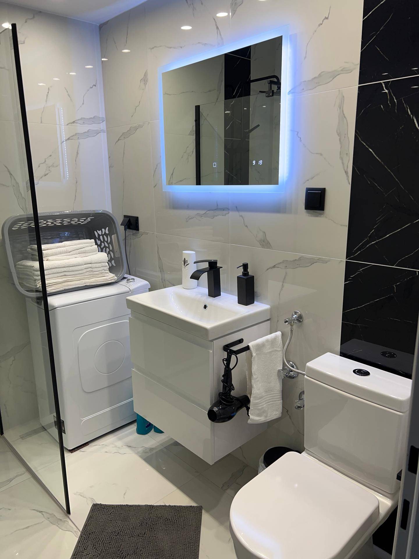 Stilvolles Badezimmer mit beleuchtetem Spiegel, modernem Waschbecken und Wand-WC auf Marmorfliesen.
