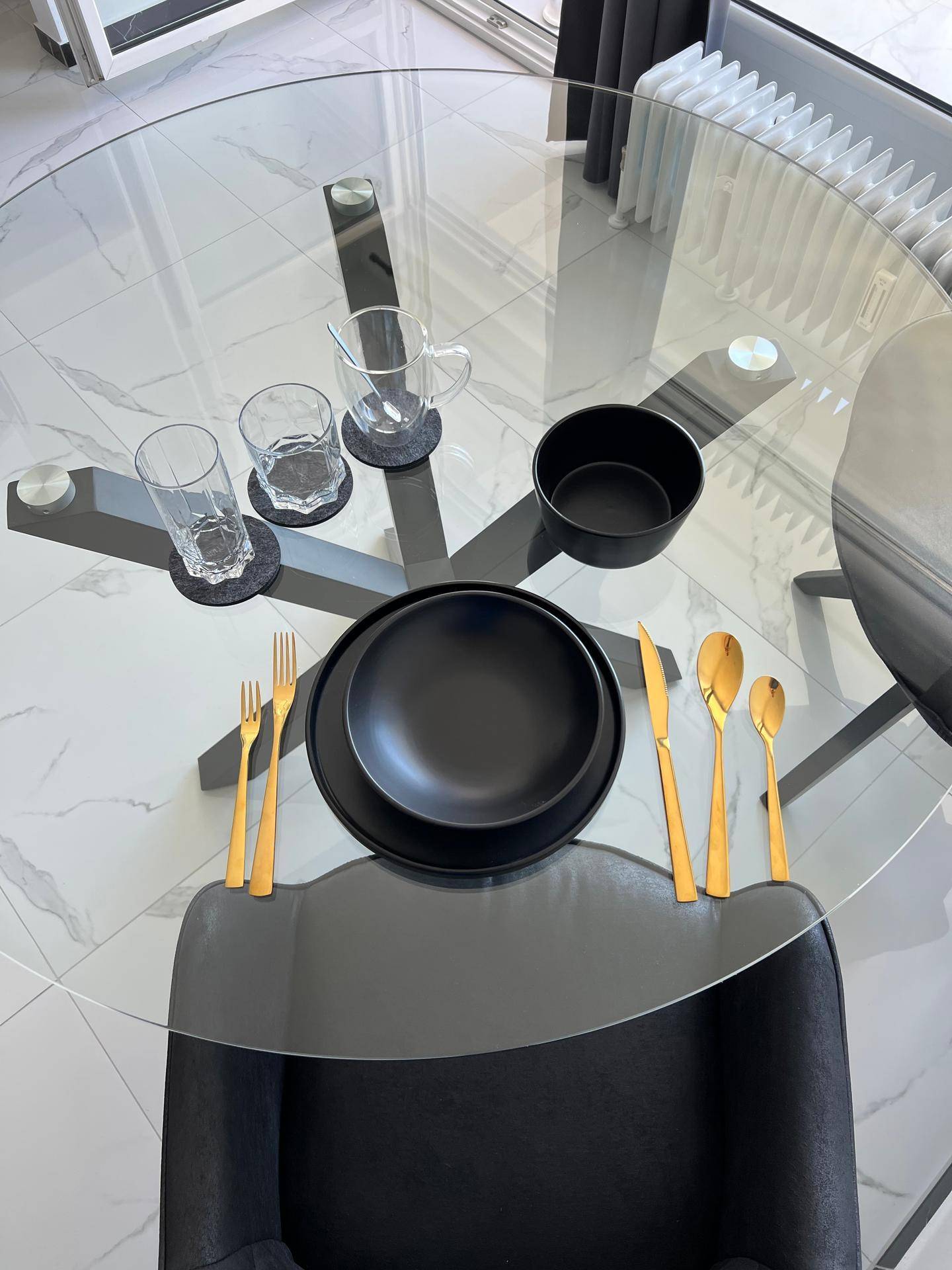 Modern gedeckter Glastisch mit schwarzen Tellern und goldenem Besteck.