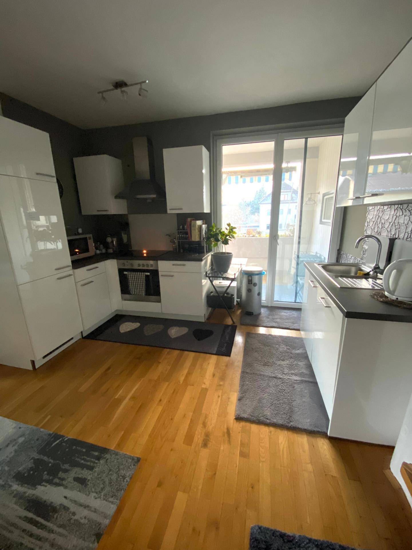 Moderne Küche mit weißen Schränken, dunkler Arbeitsplatte und Zugang zu einem sonnigen Balkon.