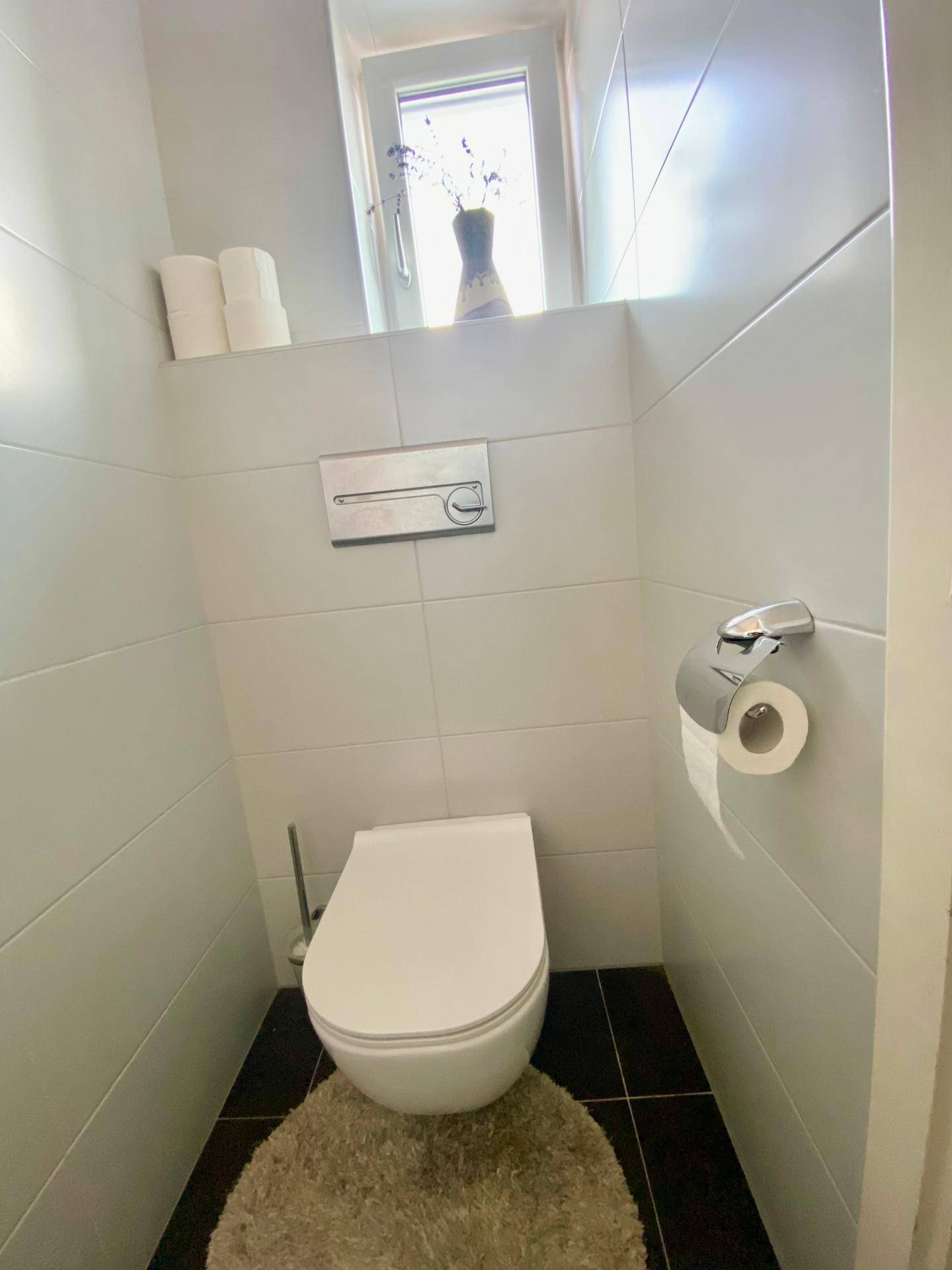 Gäste-WC mit wandhängendem WC, Fenster und dunklen Bodenfliesen, minimalistisch gestaltet.