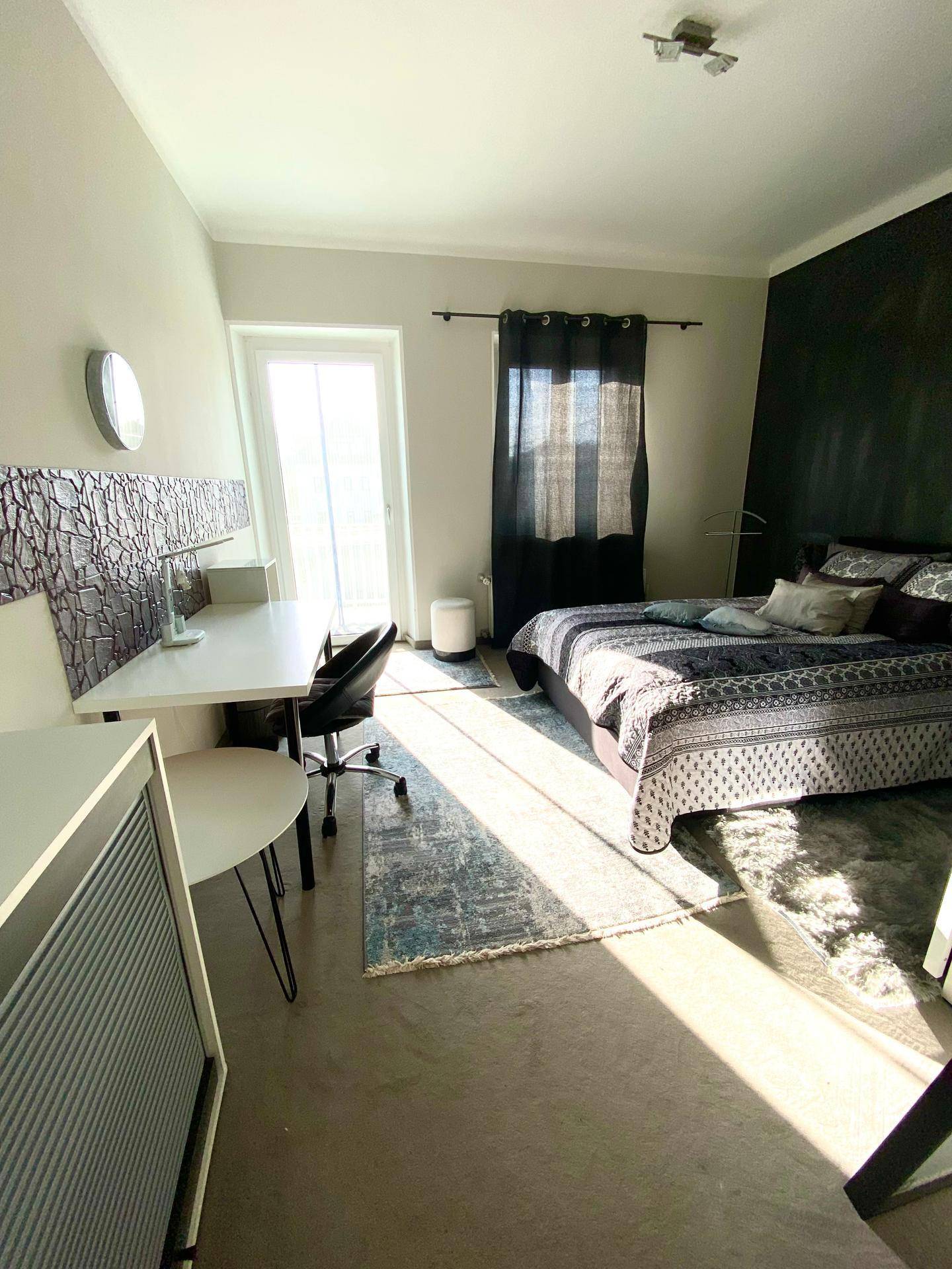 Lichtdurchflutetes Schlafzimmer mit Bett, Schreibtisch und Balkontür, die viel Tageslicht hereinlässt.