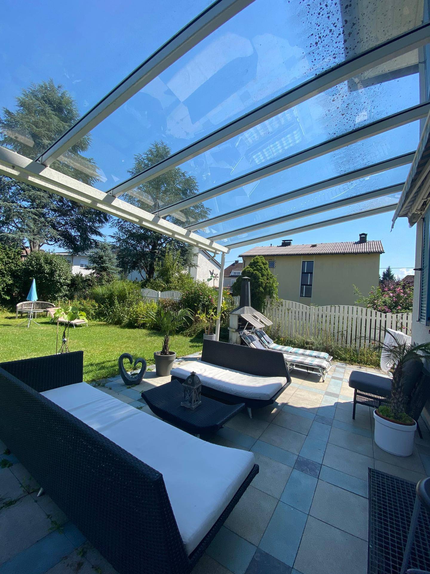 Sonnige Terrasse mit Glasdach und Lounge-Möbeln, umgeben von üppigem Grün.