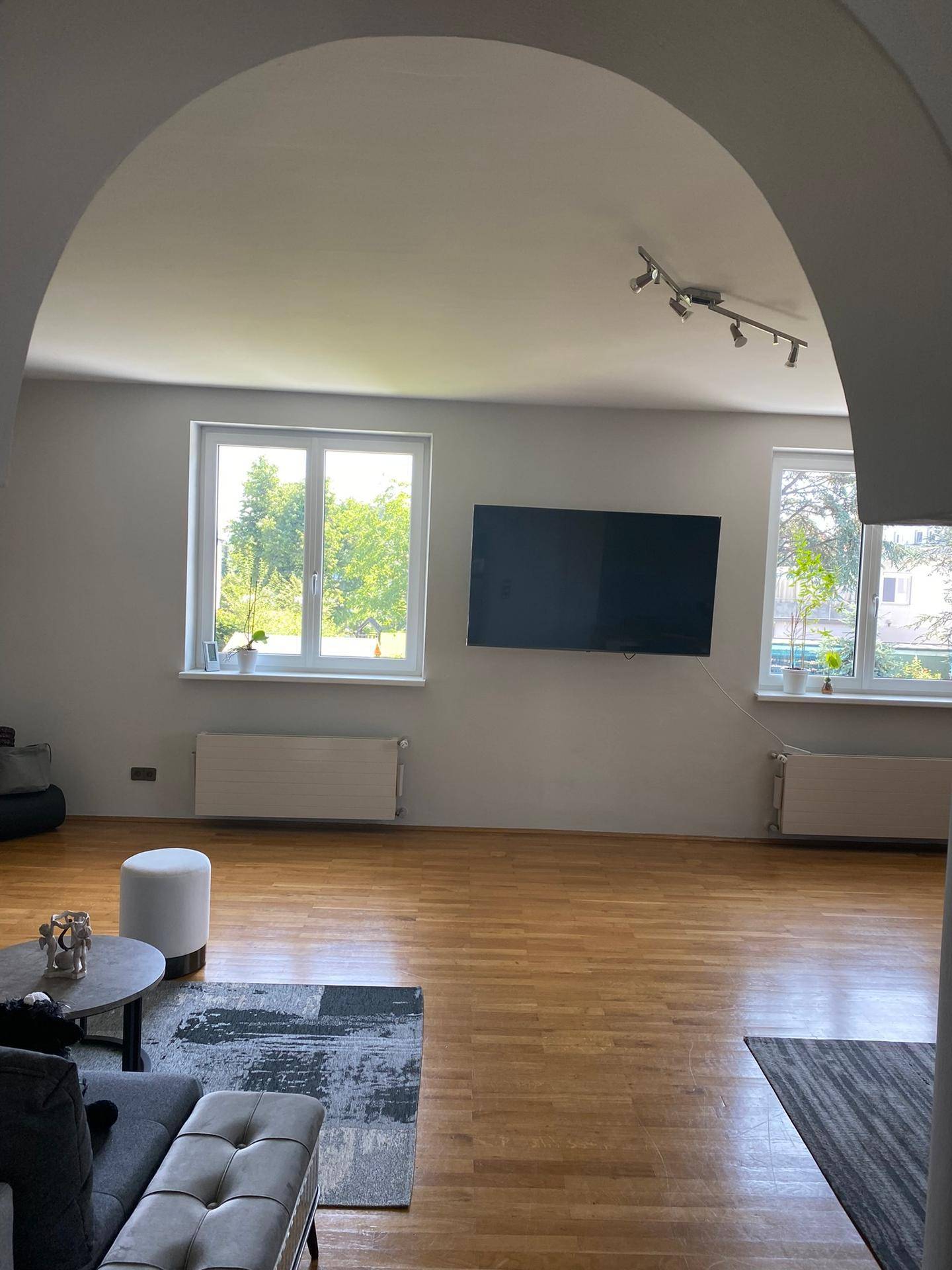 Geräumiges Wohnzimmer mit Holzboden, großen Fenstern und Wandfernseher.