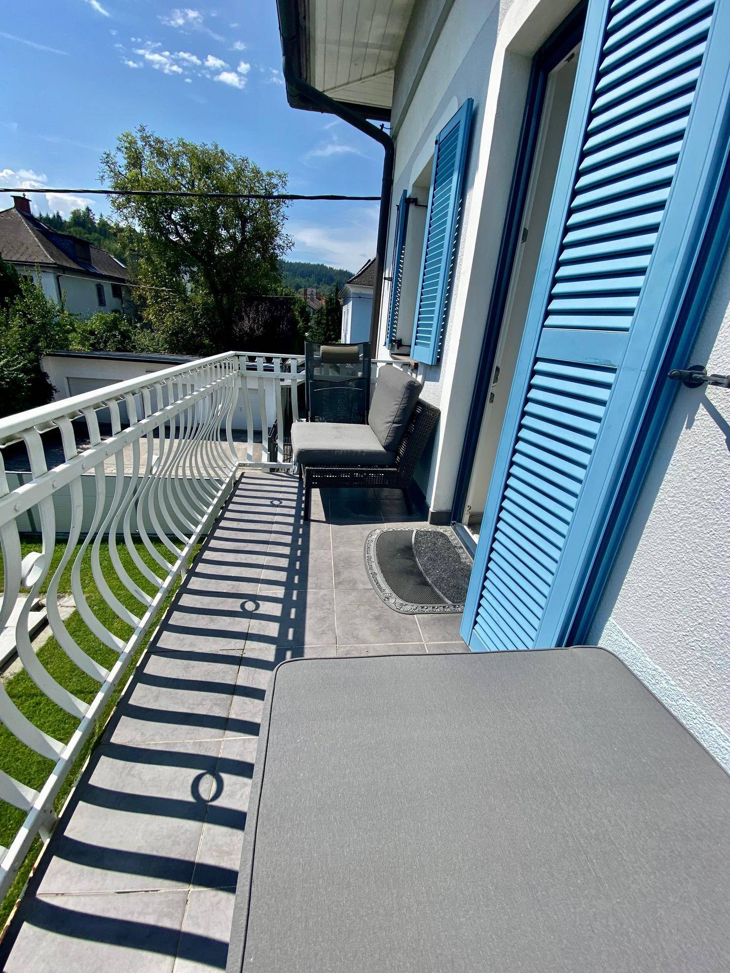 Schmaler Balkon mit blauen Fensterläden, weißem Geländer und Sitzgelegenheiten im Freien.