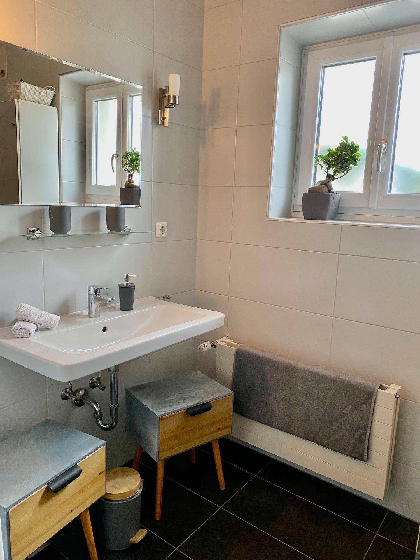 Modernes Badezimmer mit Waschbecken, Spiegel, Fenster und dunklen Bodenfliesen.