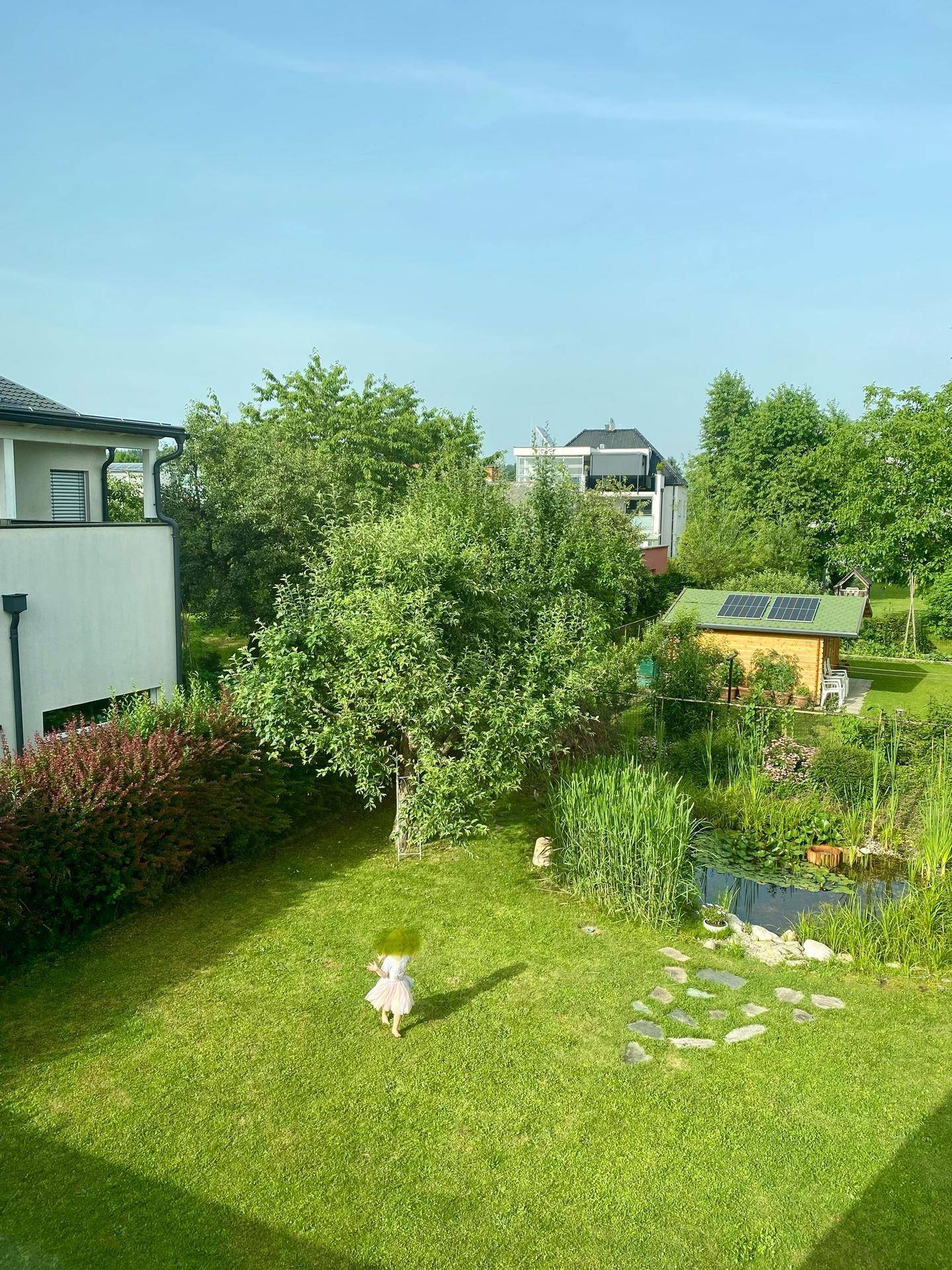 Gepflegter grüner Garten mit Rasenfläche, Teich und Gartenhaus mit Solaranlage.