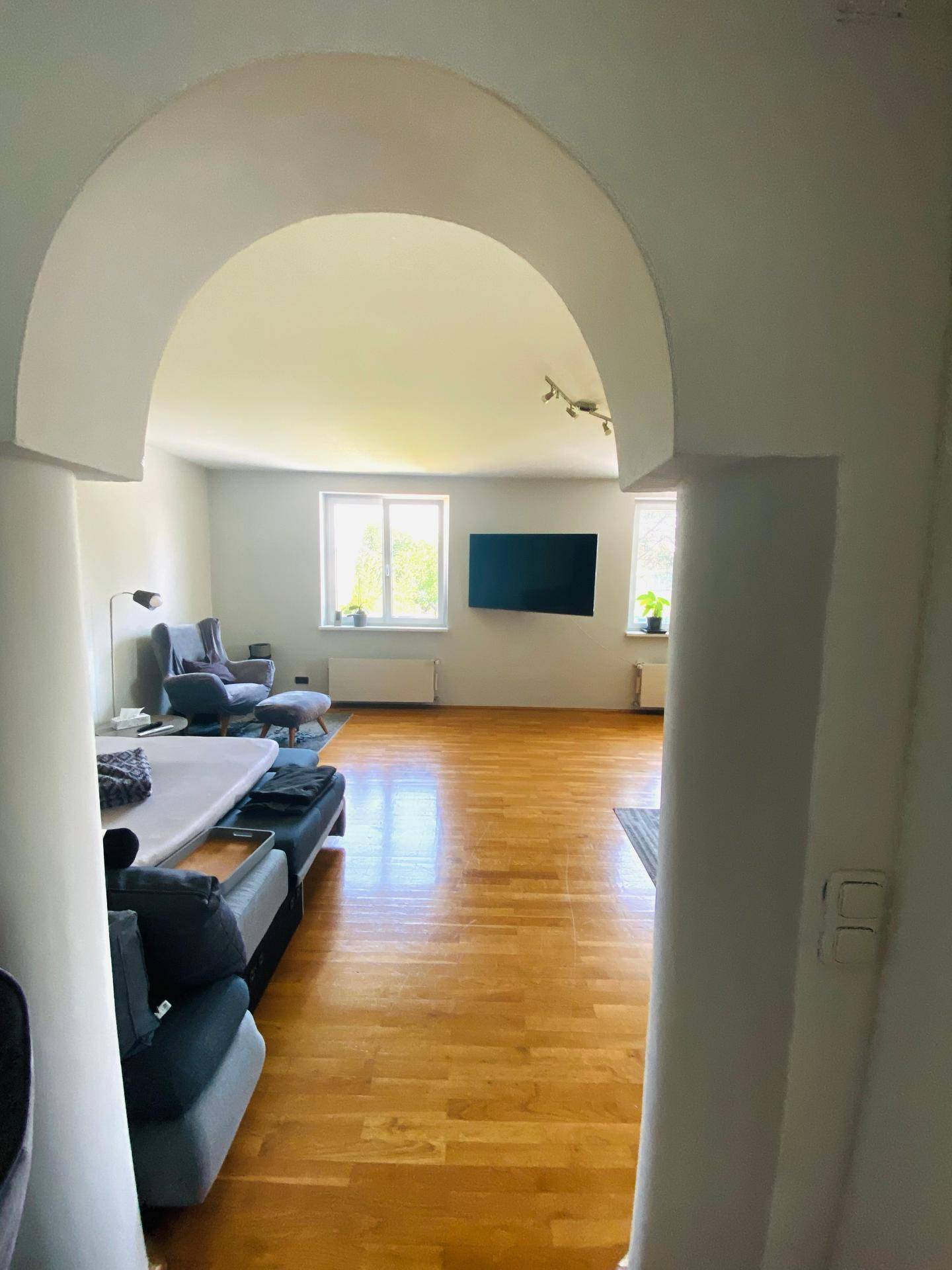 Heller Wohnbereich mit großem Fenster, Holzboden und einem an der Wand montierten Fernseher.