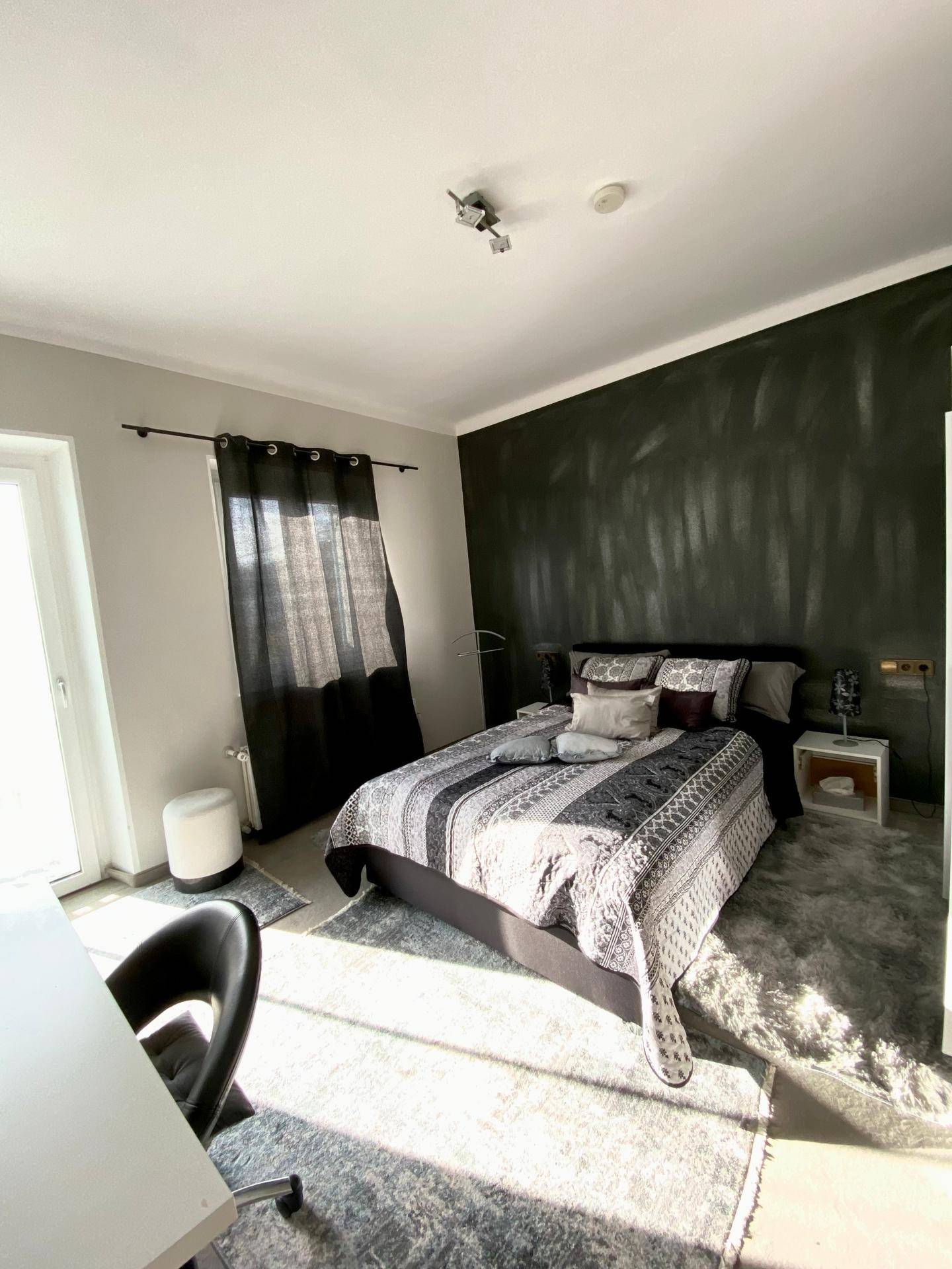 Helles Schlafzimmer mit modernem Bett, dunkler Akzentwand und Zugang zu einem Balkon.