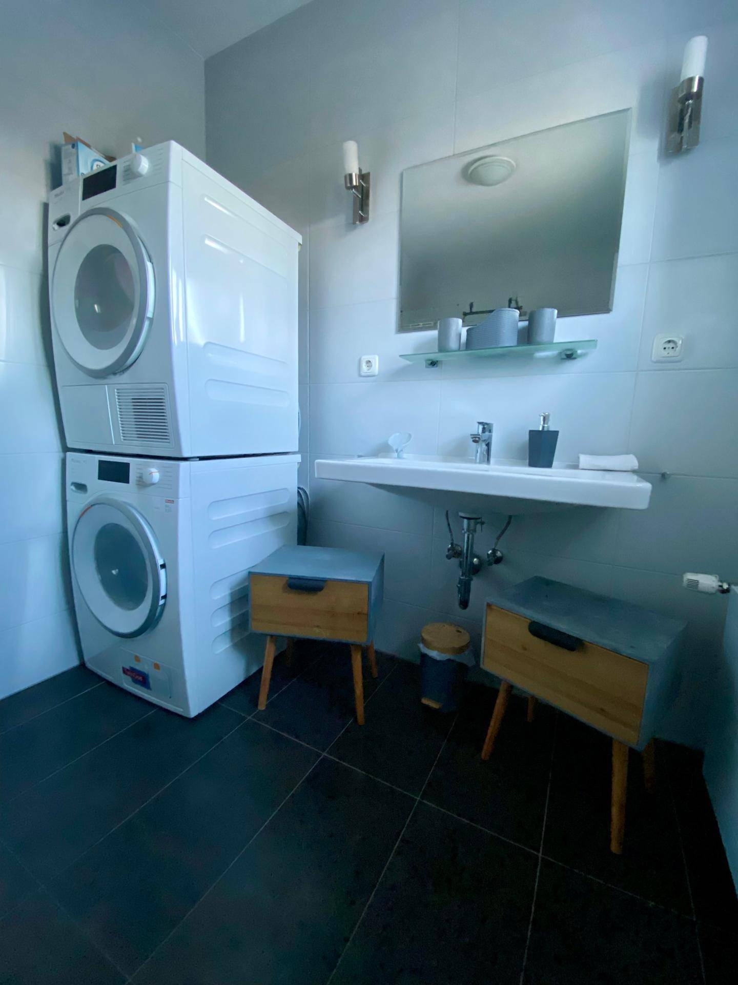 Funktionales Badezimmer mit Waschbecken, Spiegel und gestapeltem Waschtrockner, hell gefliest.