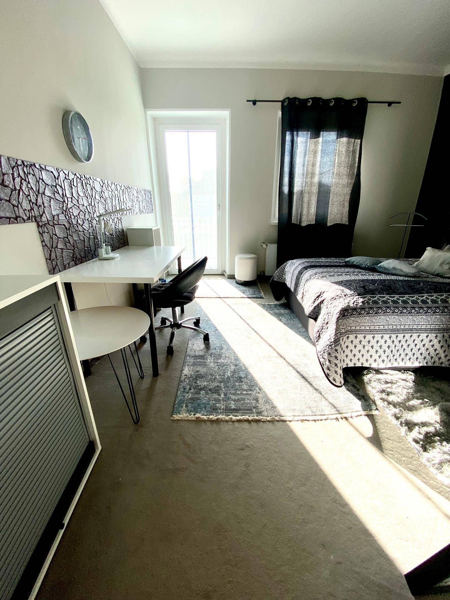 Helles Schlafzimmer mit Bett, Schreibtisch und Zugang zu einem Balkon, durchflutet von Sonnenlicht.