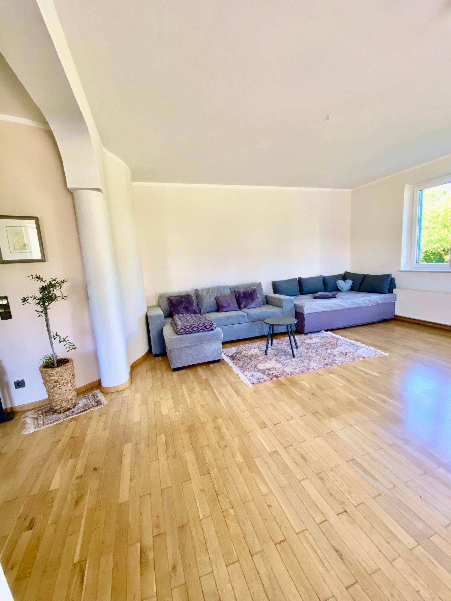 Geräumiges Wohnzimmer mit Holzboden, großem Sofa und einem Fenster mit Blick ins Grüne.