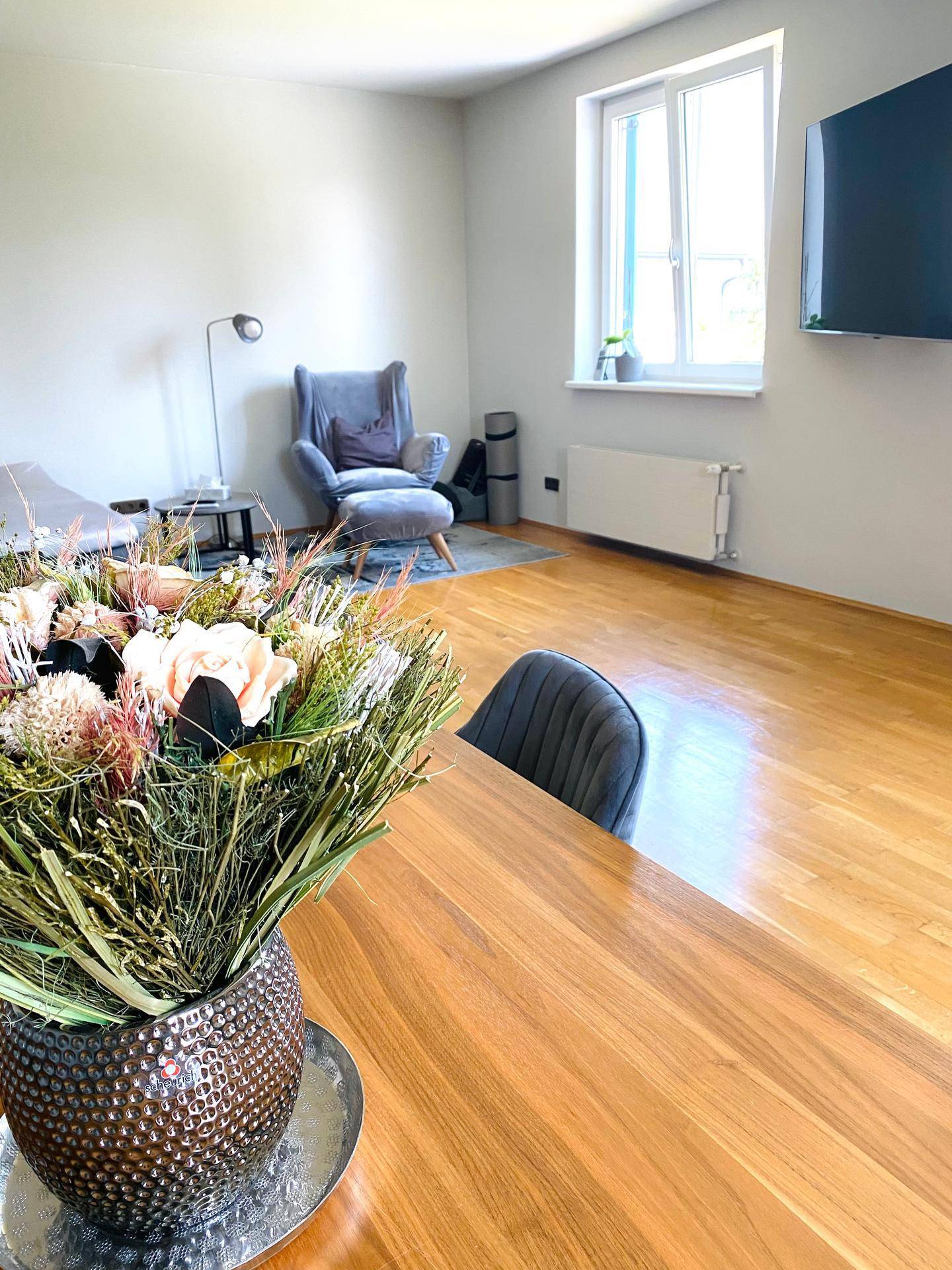 Lichtdurchflutetes Wohnzimmer mit Holzboden, großem Fenster und einem Blumenarrangement auf dem Tisch.
