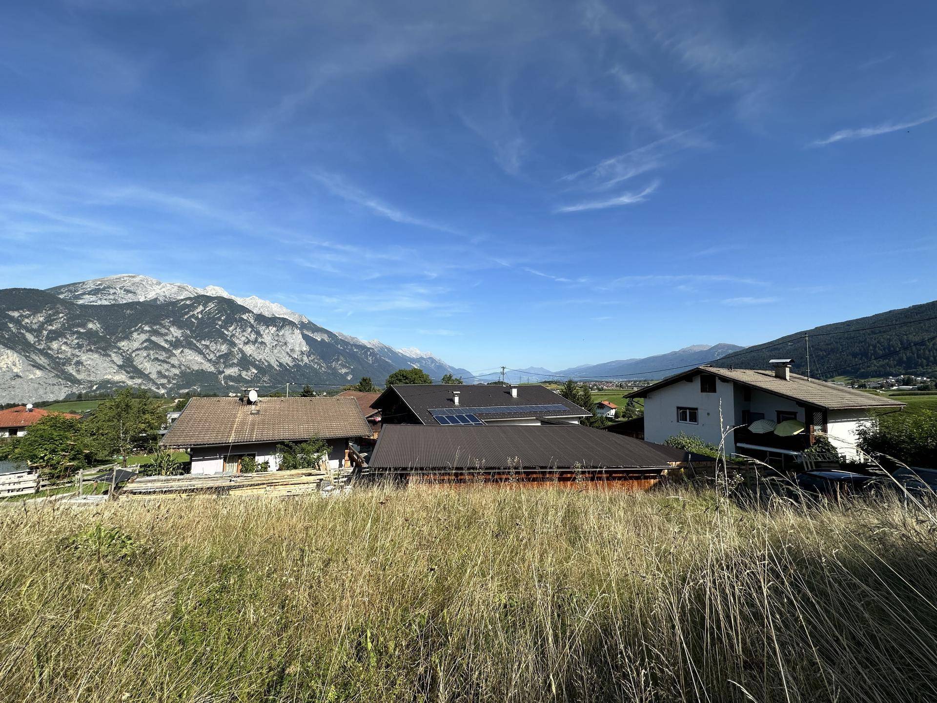 Panoramablick auf Wohnhäuser mit Solaranlagen und die umliegende Berglandschaft unter blauem Himmel.