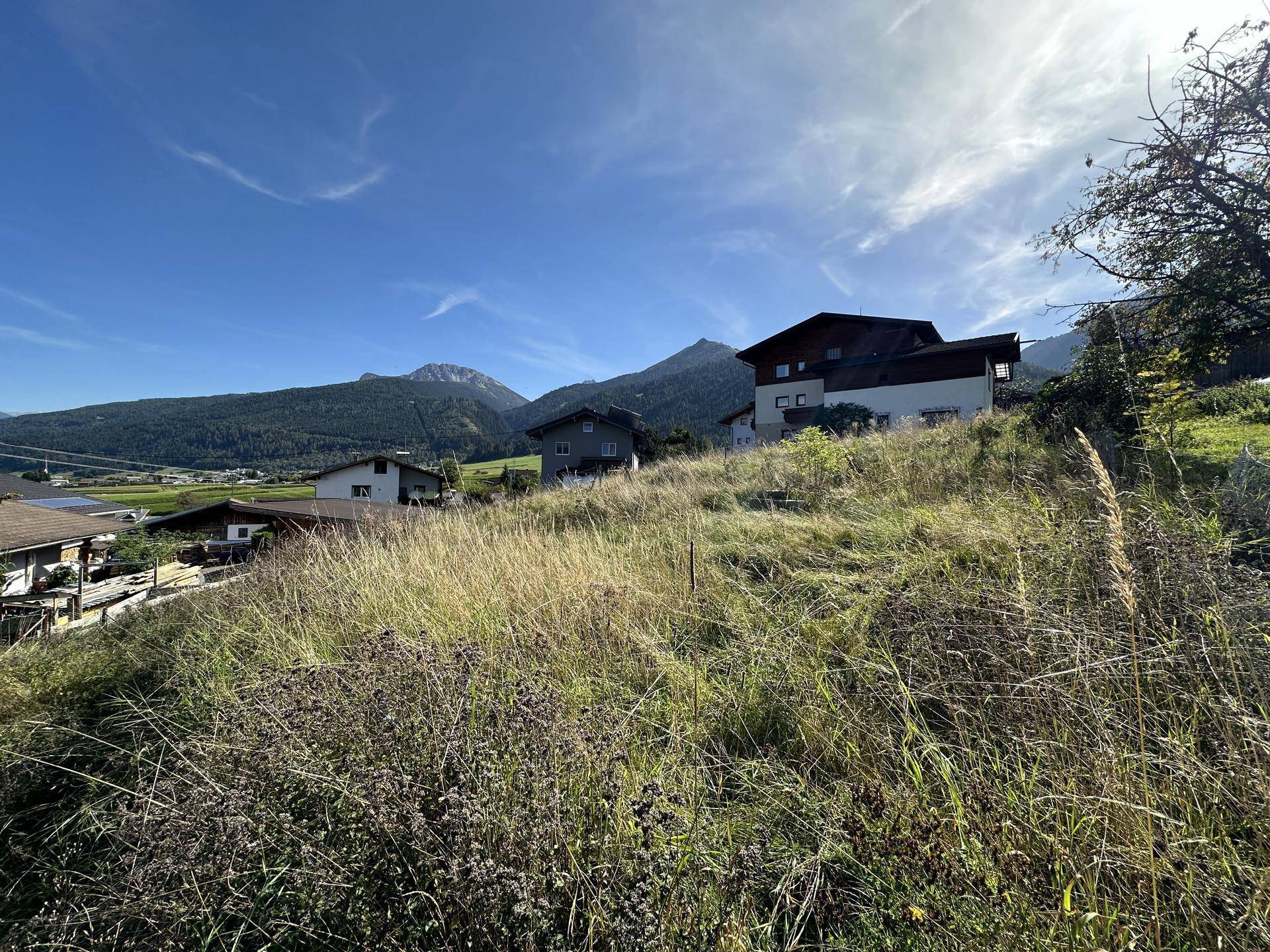 Sonniger Blick auf eine hügelige Landschaft mit Wohnhäusern und bewaldeten Bergen am Horizont.