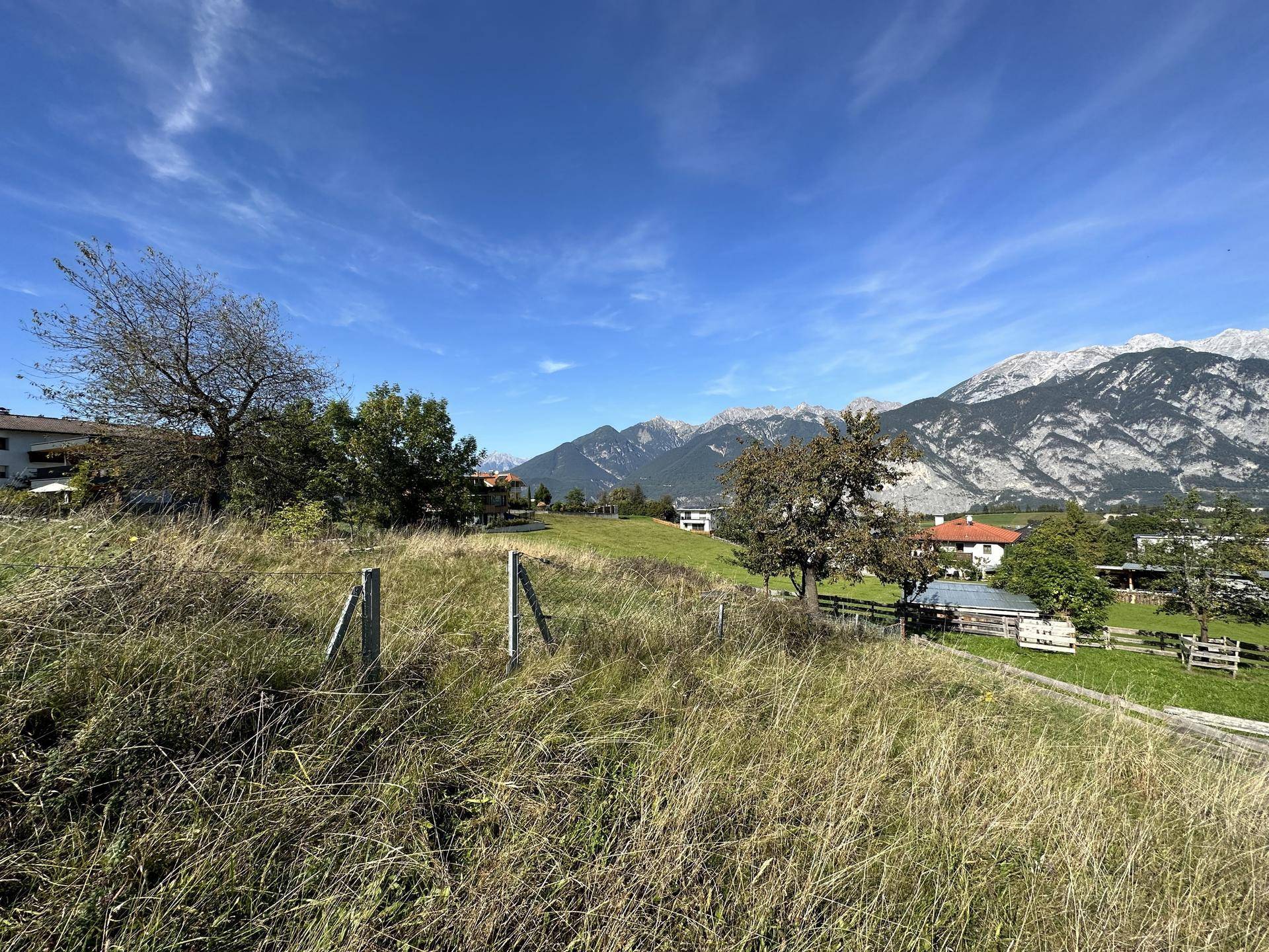 Weitläufige Wiesenlandschaft mit vereinzelten Bäumen, Häusern und einer majestätischen Bergkette.