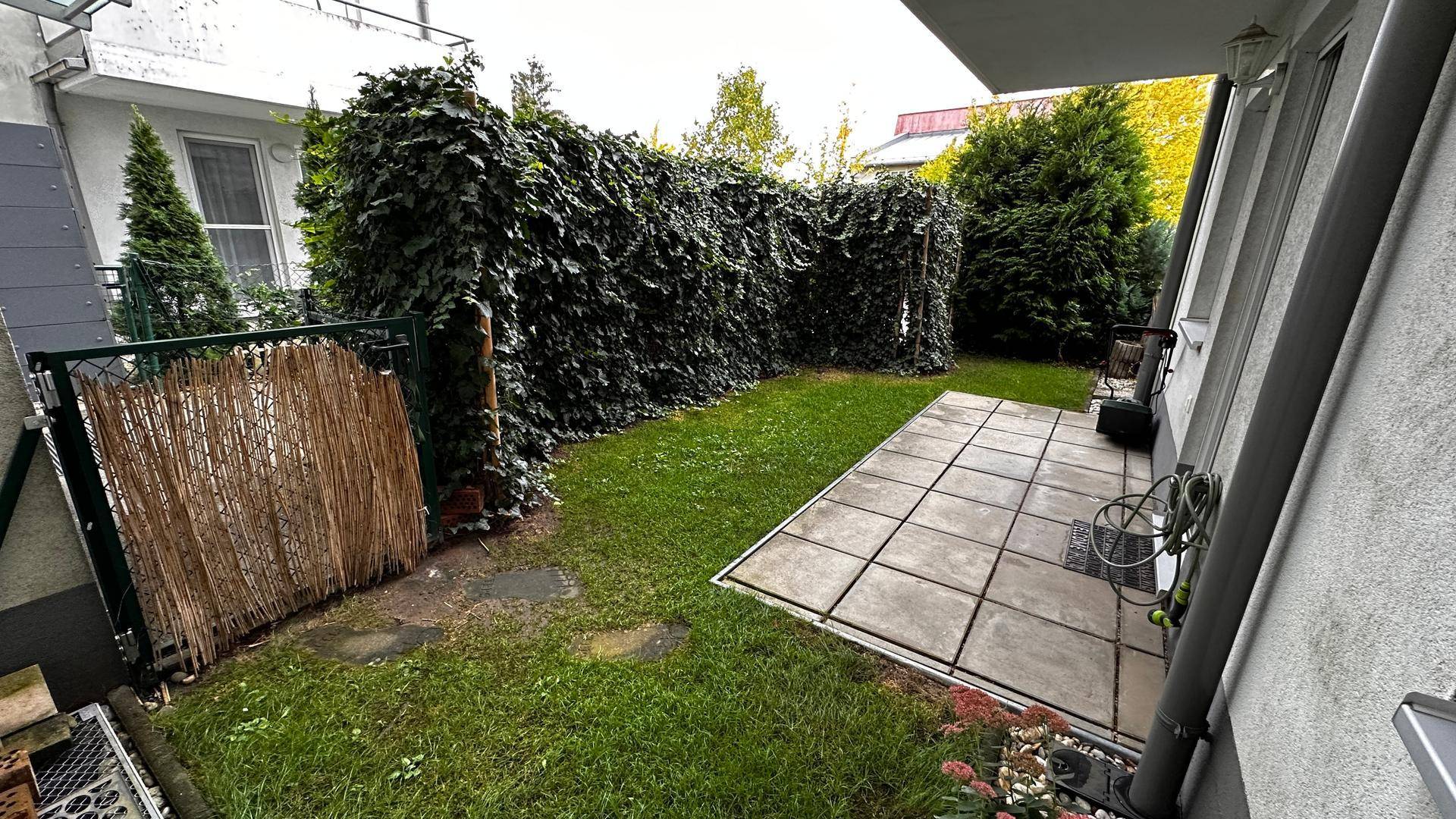 Privater Gartenbereich mit gepflasterter Terrasse, umgeben von einer hohen grünen Hecke.