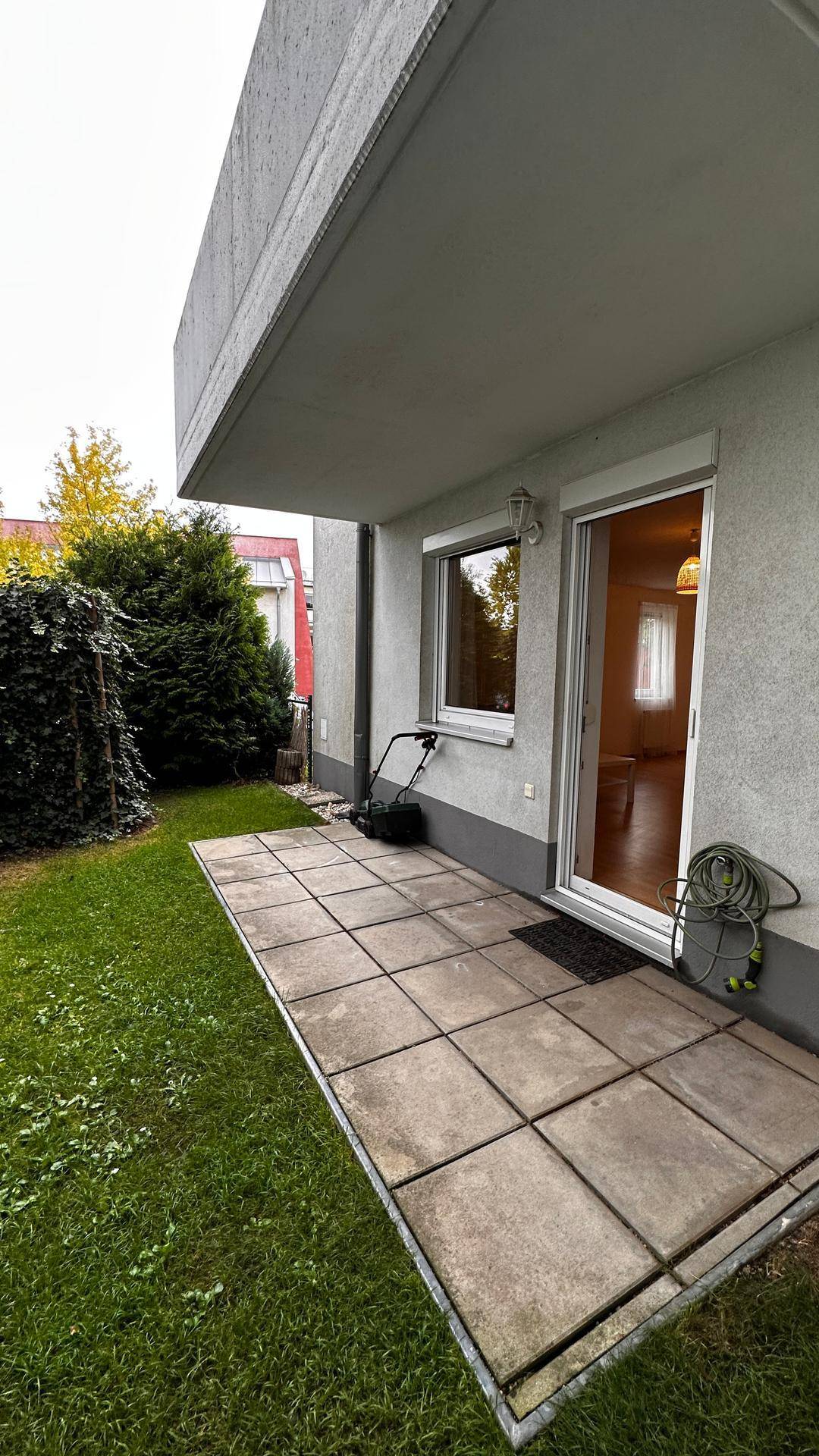 Kleine gepflasterte Terrasse neben dem Gebäude mit Rasenfläche und Hecke.