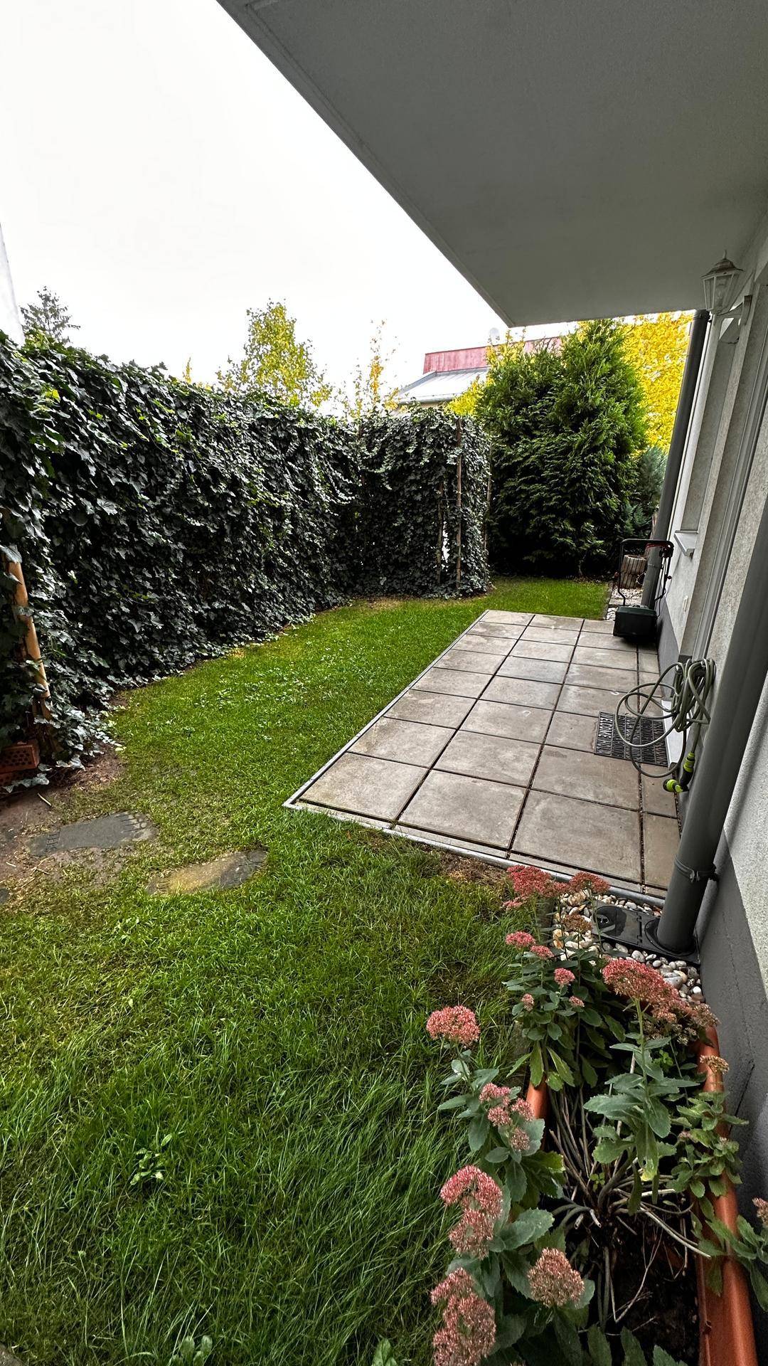 Grüne Gartenfläche mit kleiner Terrasse und Blumenbeeten am Rand.