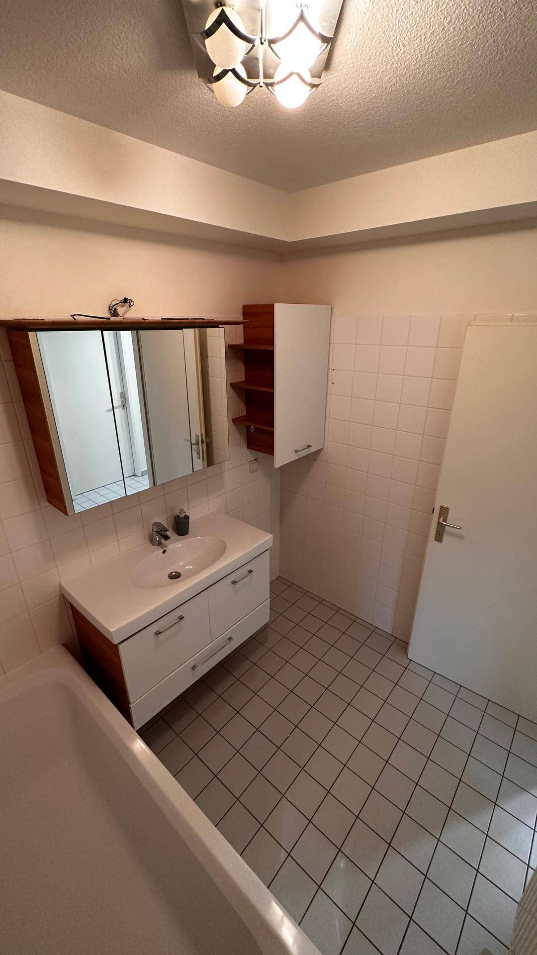 Badezimmer mit Waschtisch, Spiegelschrank und Zugangstür, in hellen Fliesen gehalten.