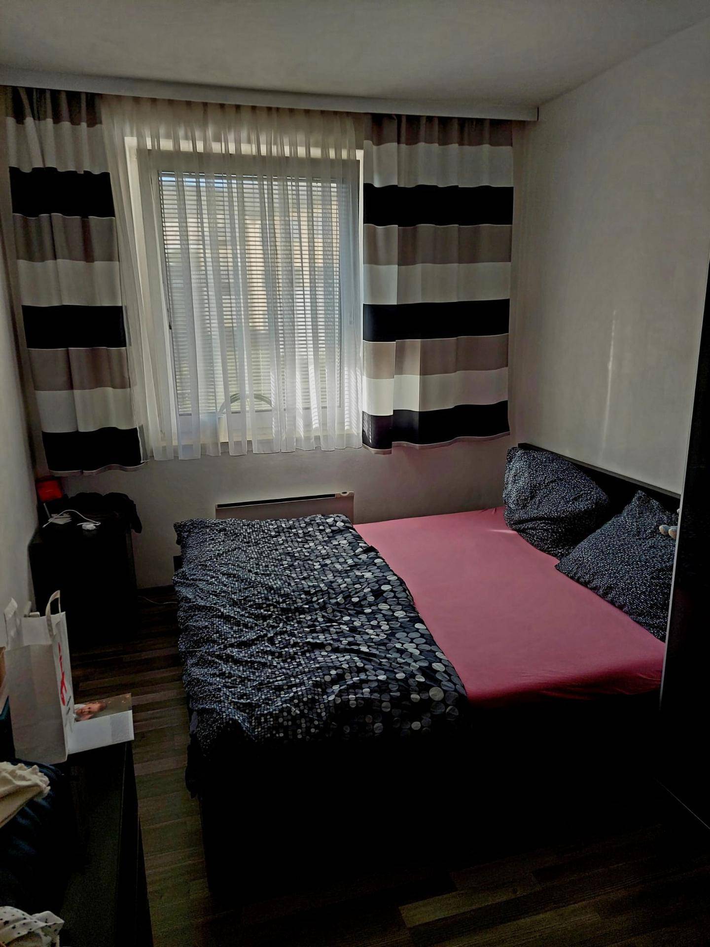 Schlafzimmer mit Bett, Fenster mit Jalousien und Vorhängen, ausgestattet mit dunklem Laminatboden.