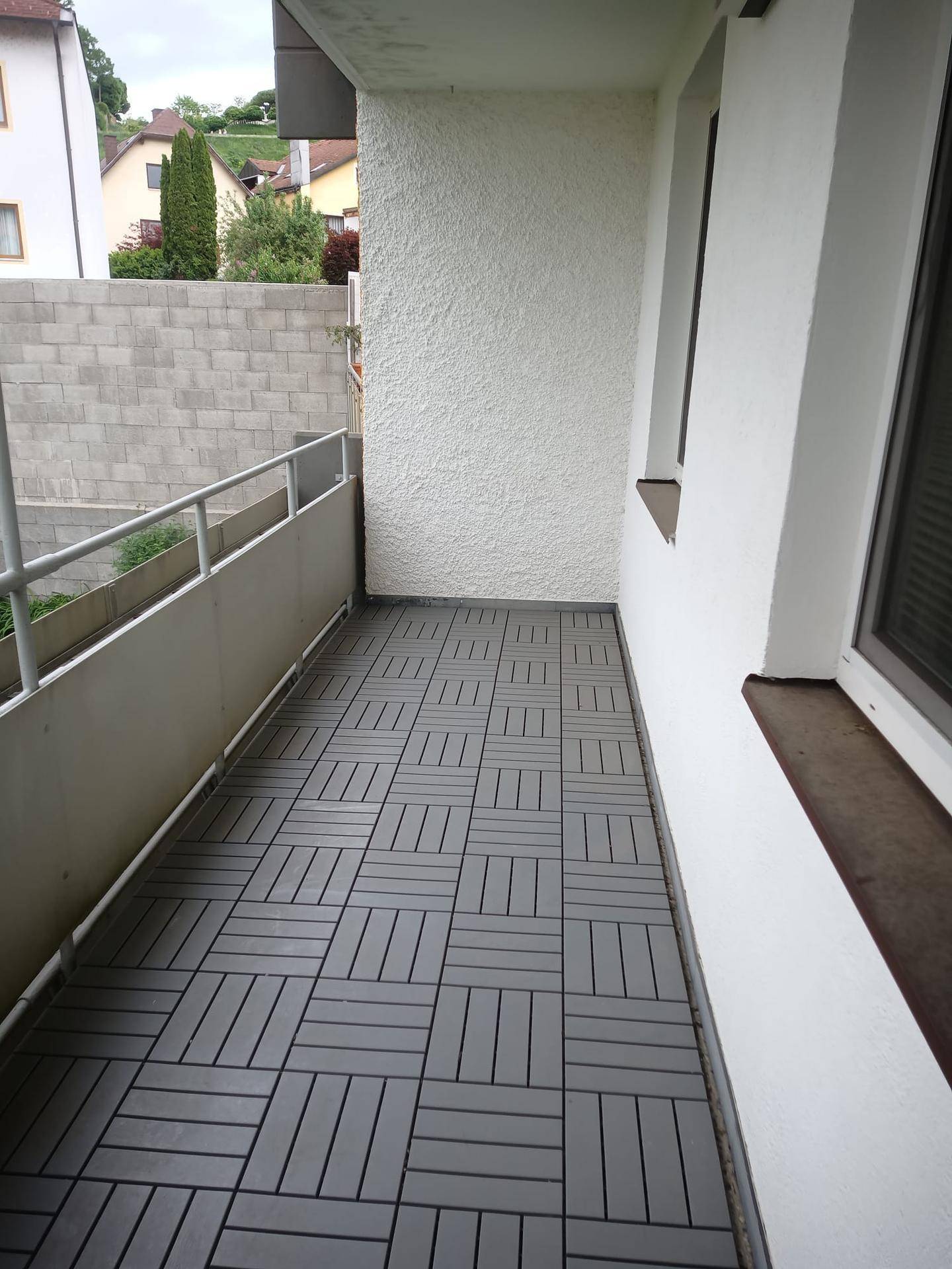 Balkon mit Metallgeländer und grauen Bodenfliesen, bietet einen Blick auf eine Mauer und Grünflächen.
