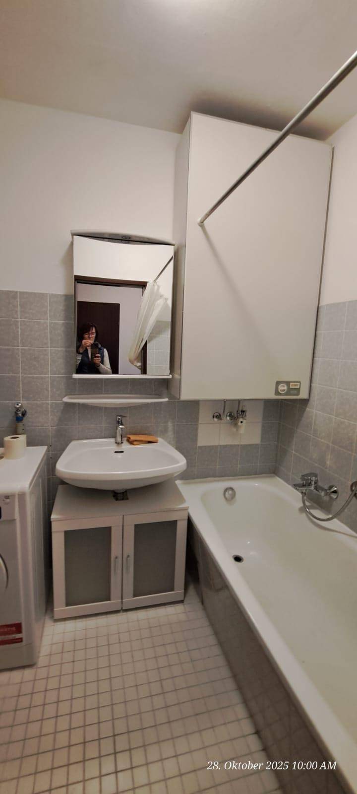 Badezimmer mit Badewanne, Waschbecken, Spiegel und Waschmaschine, gefliest in Grau und Weiß.
