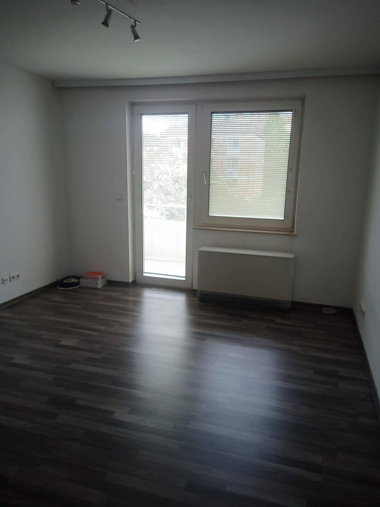Leerer Raum mit dunklem Laminatboden und Zugang zu einem Balkon durch eine Fenstertür.