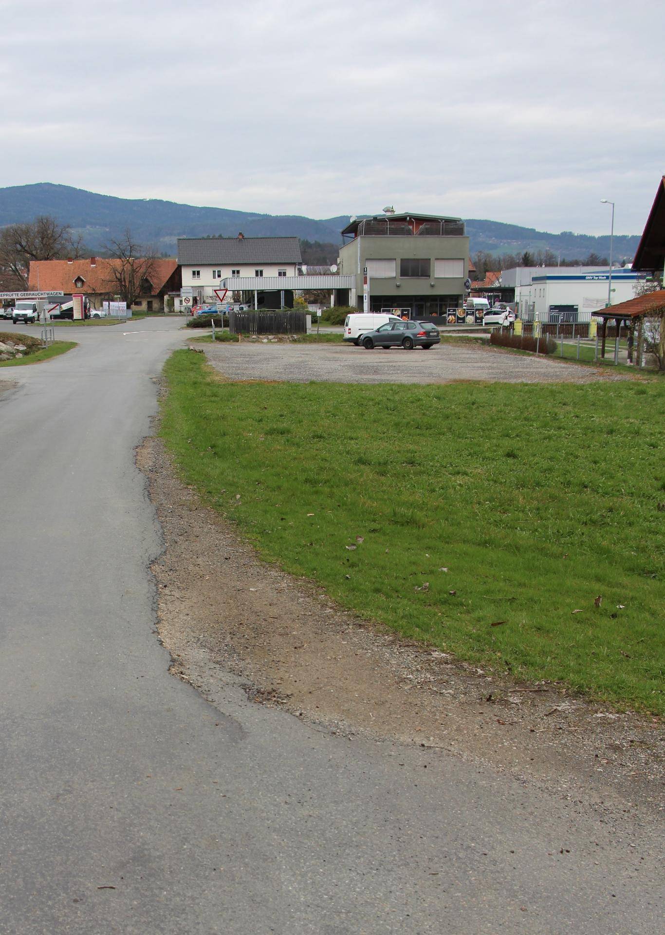 Blick auf eine ländliche Straße mit Gebäuden, Grünflächen und Bergen im Hintergrund.