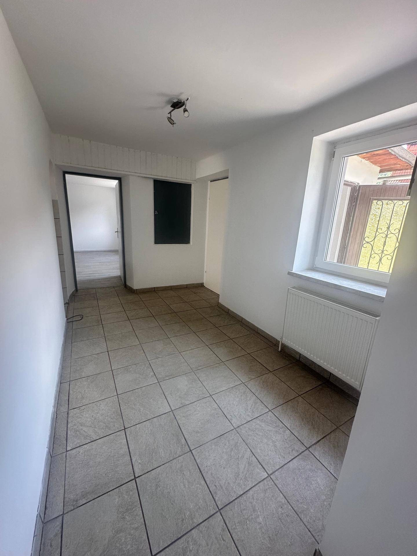 Heller Flur mit grauen Fliesen, Heizkörper unter dem Fenster und Zugang zu weiteren Räumen.