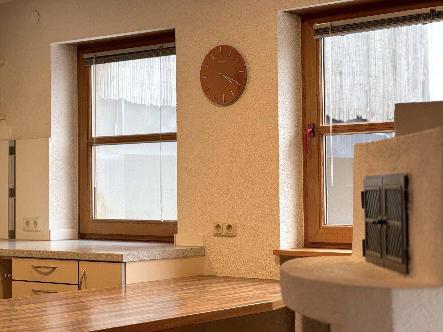 Detailansicht der Küche mit zwei Fenstern, einer Wanduhr und dem traditionellen Kachelofen.