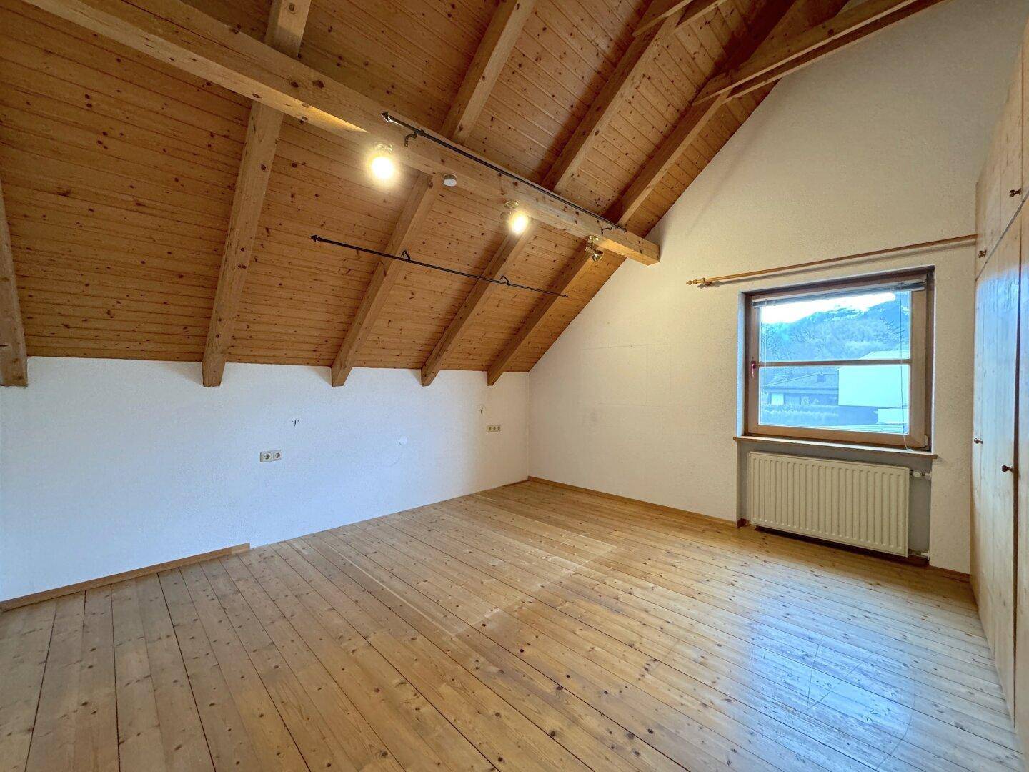 Helles Schlafzimmer im Dachgeschoss mit Holzboden, sichtbaren Holzbalken und einem Fenster.