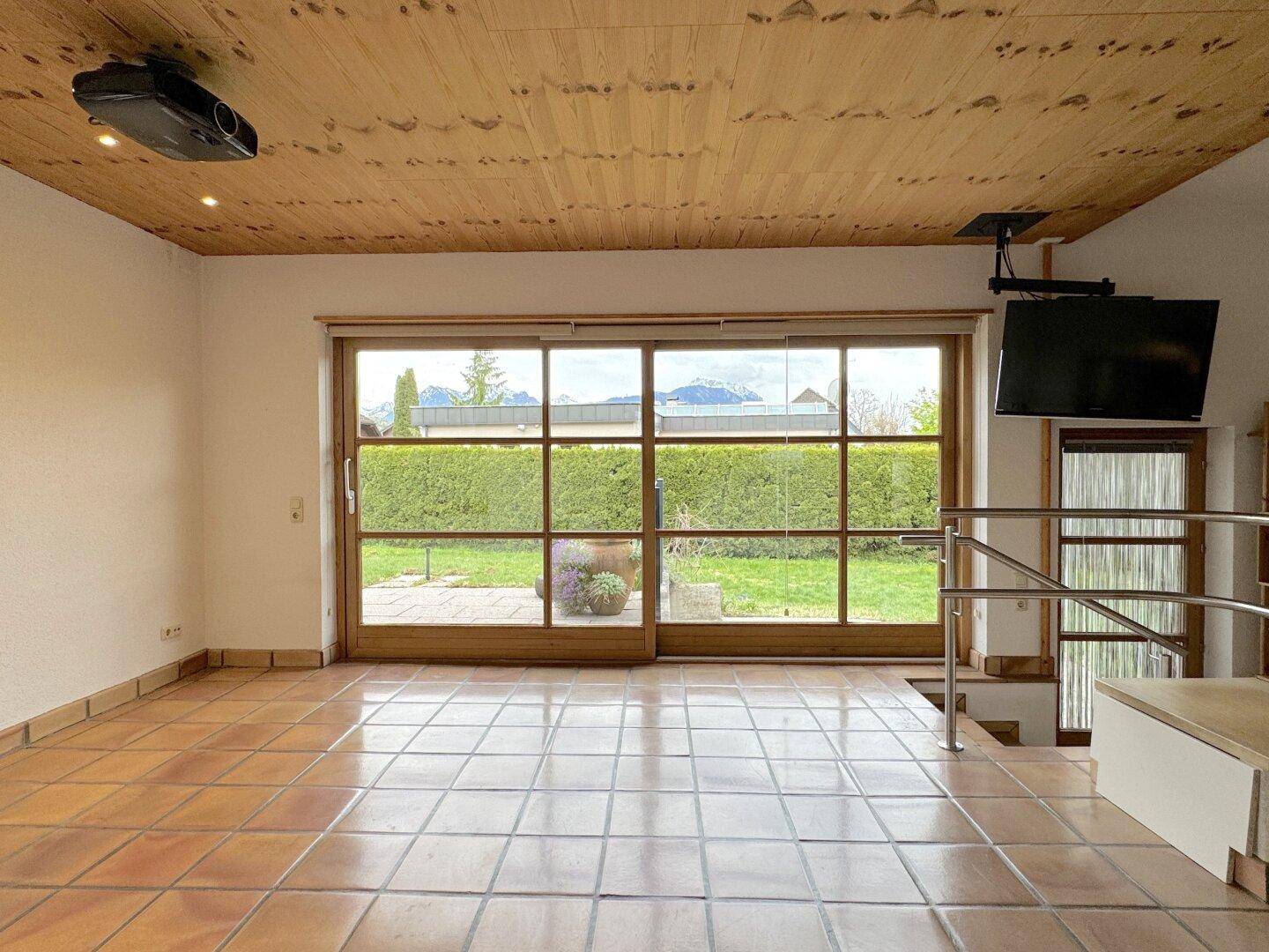 Heller Wohnbereich mit Fliesenboden, Holzdecke und großen Fenstern mit Blick auf den Garten und die Berge.