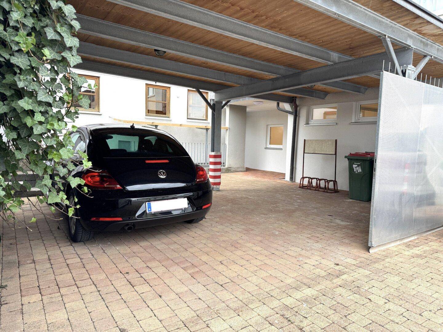 Überdachter Carport mit gepflastertem Boden und einem geparkten Auto, umgeben von Grün.