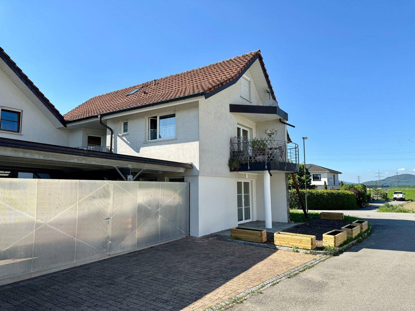 Ansicht des Hauses mit Carport und einem kleinen Balkon, umgeben von viel Grün und einem weiten Blick.