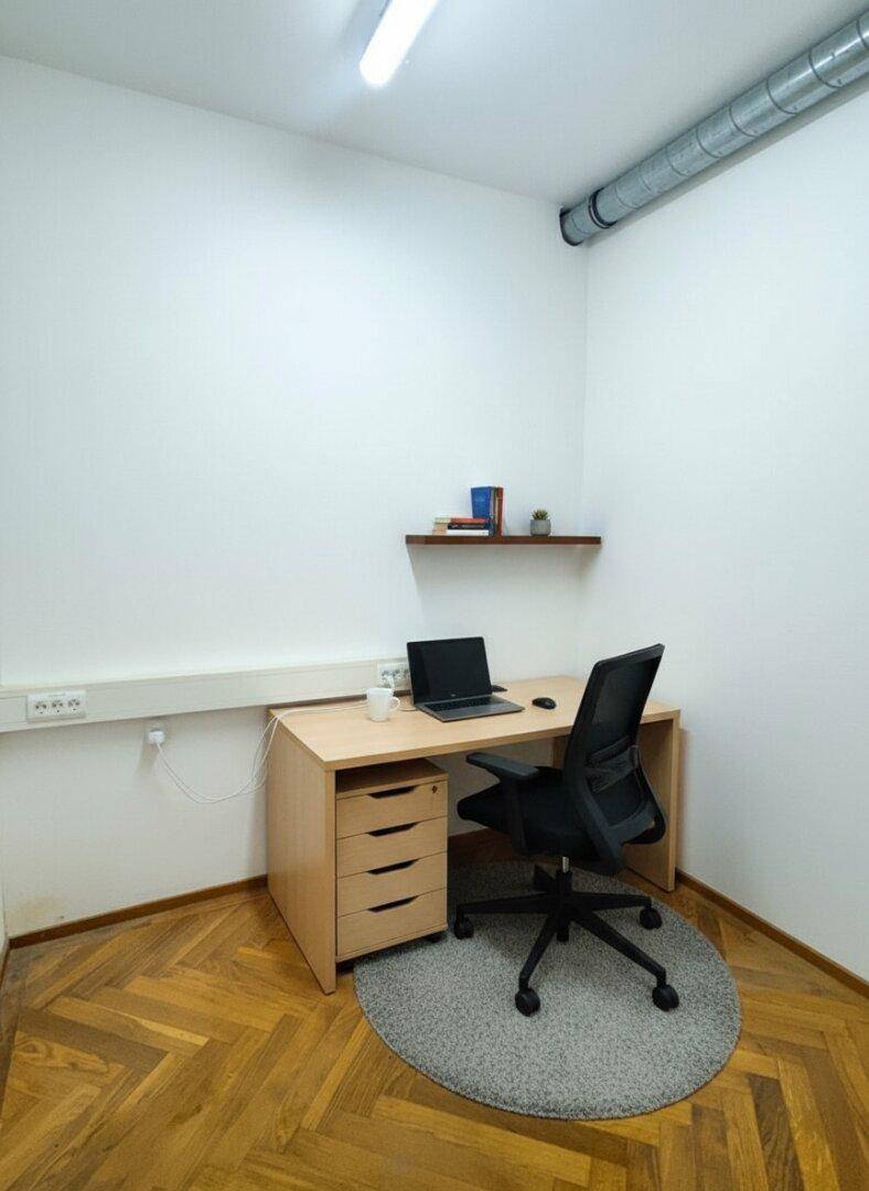 Kleines, helles Büro mit einem Schreibtisch, Bürostuhl und einem Regal an der Wand.