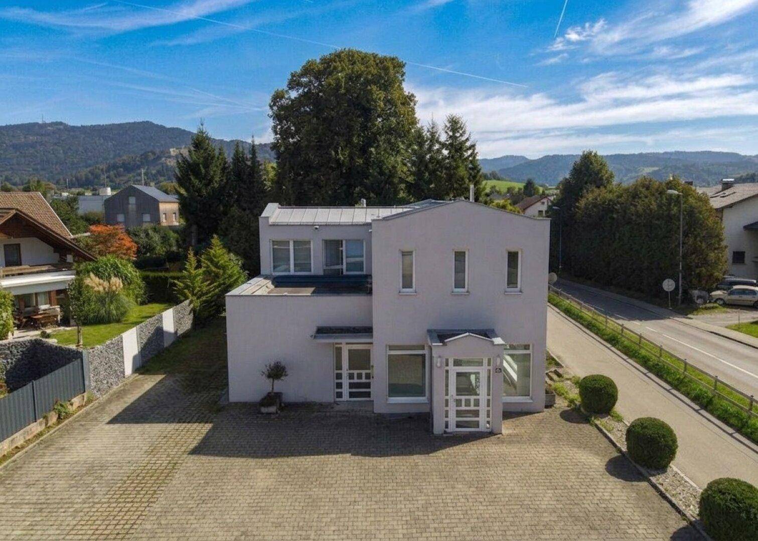 Modernes Gebäude mit heller Fassade und gepflastertem Vorplatz, umgeben von grüner Landschaft und Bergen.