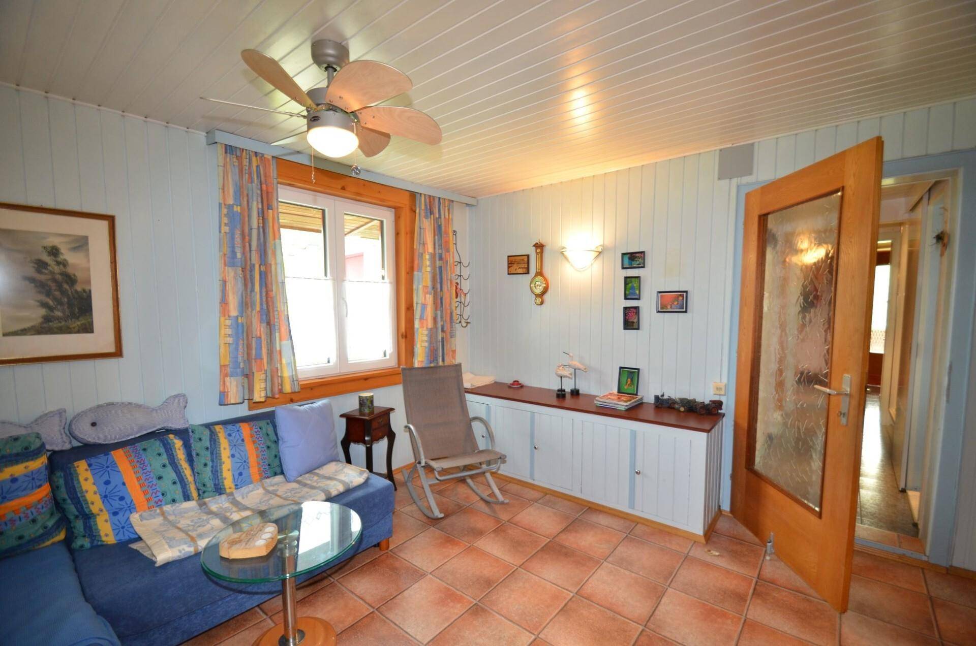 Helles Wohnzimmer mit blauem Ecksofa, Deckenventilator und Terrakottafliesen.